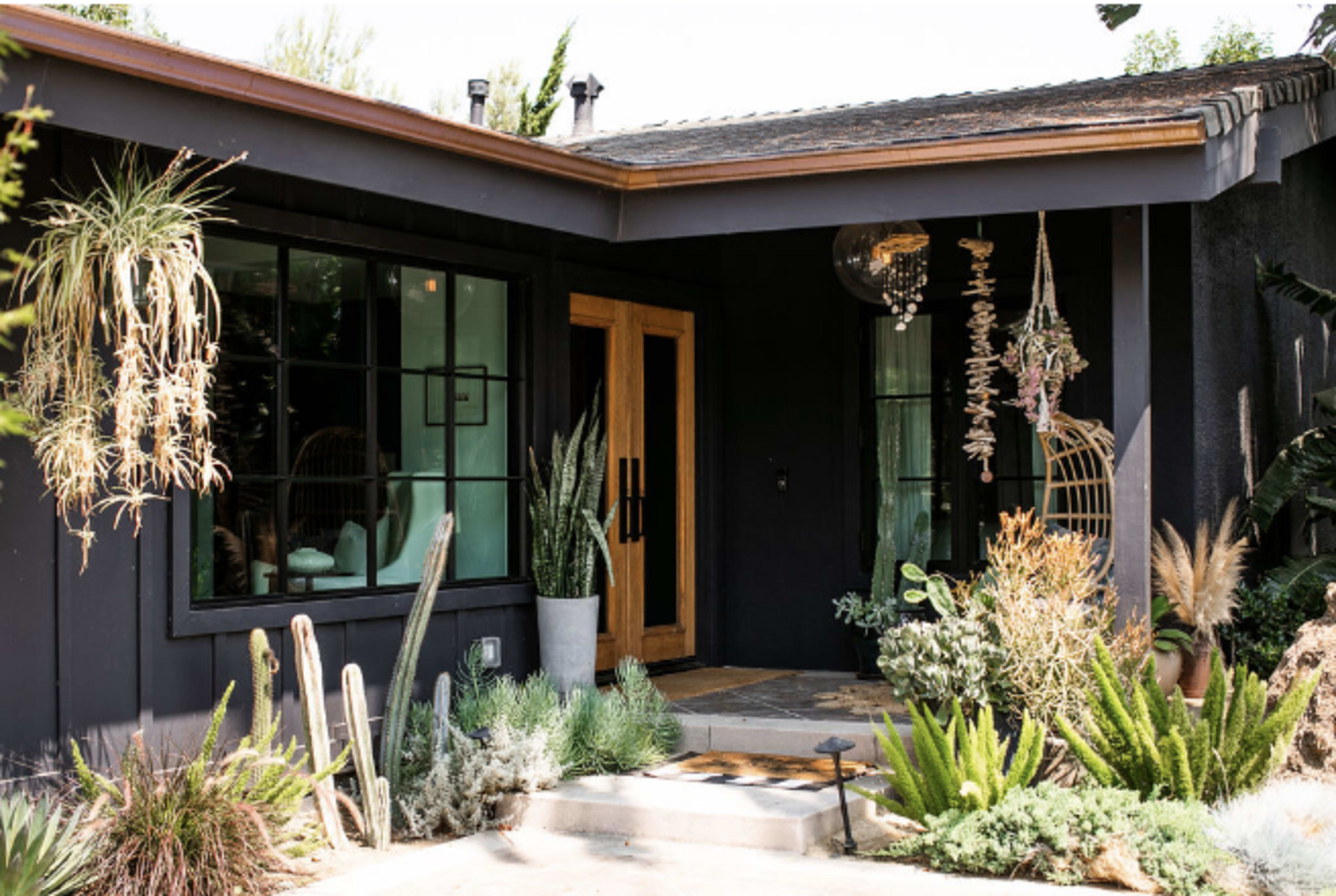 The image features a modern black house entrance with a wooden door, surrounded by various potted plants and decorative hanging elements.