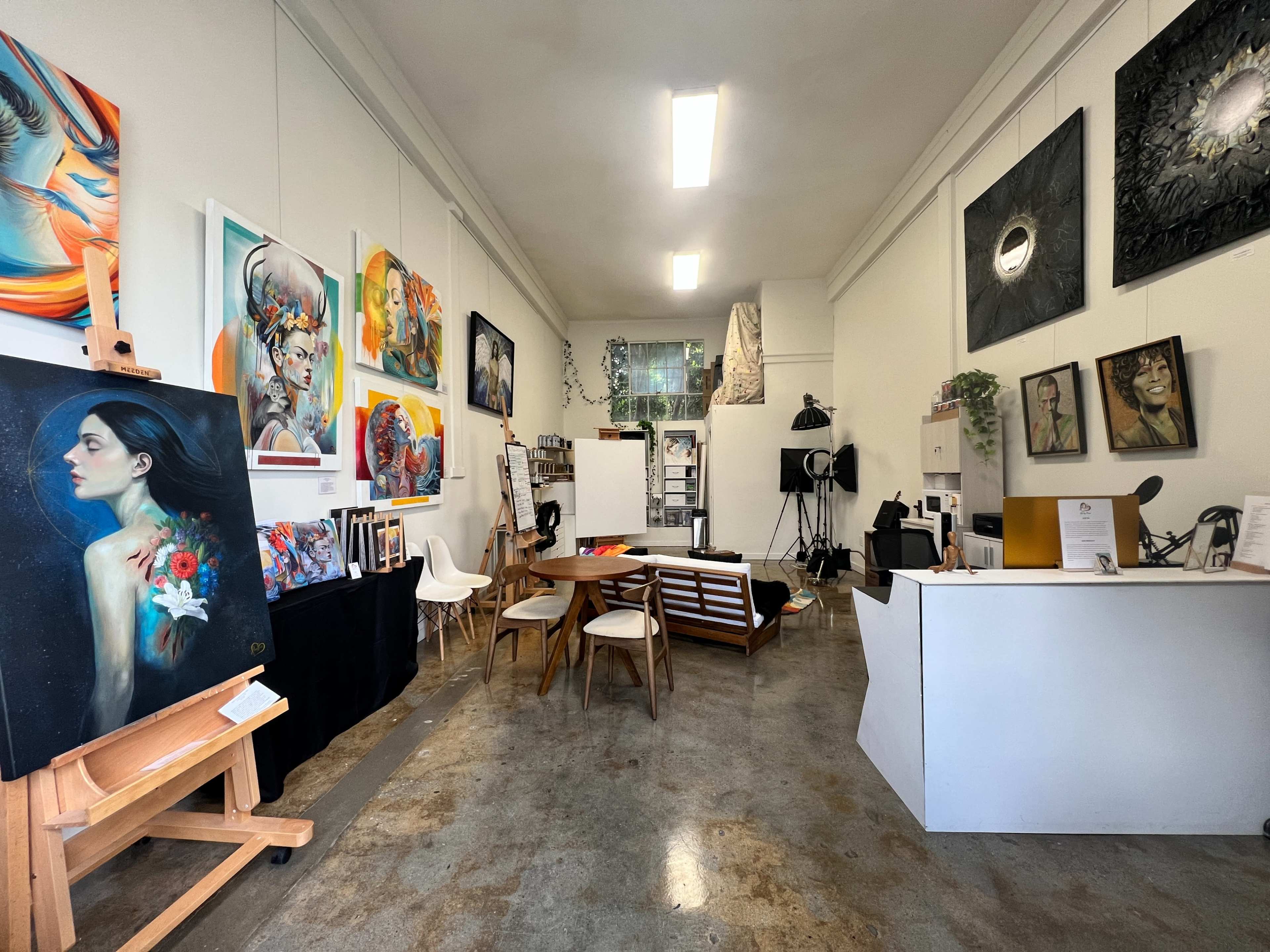 The image shows an art studio with various colorful paintings displayed on the walls, a wooden table and chairs in the center, and an easel on one side.