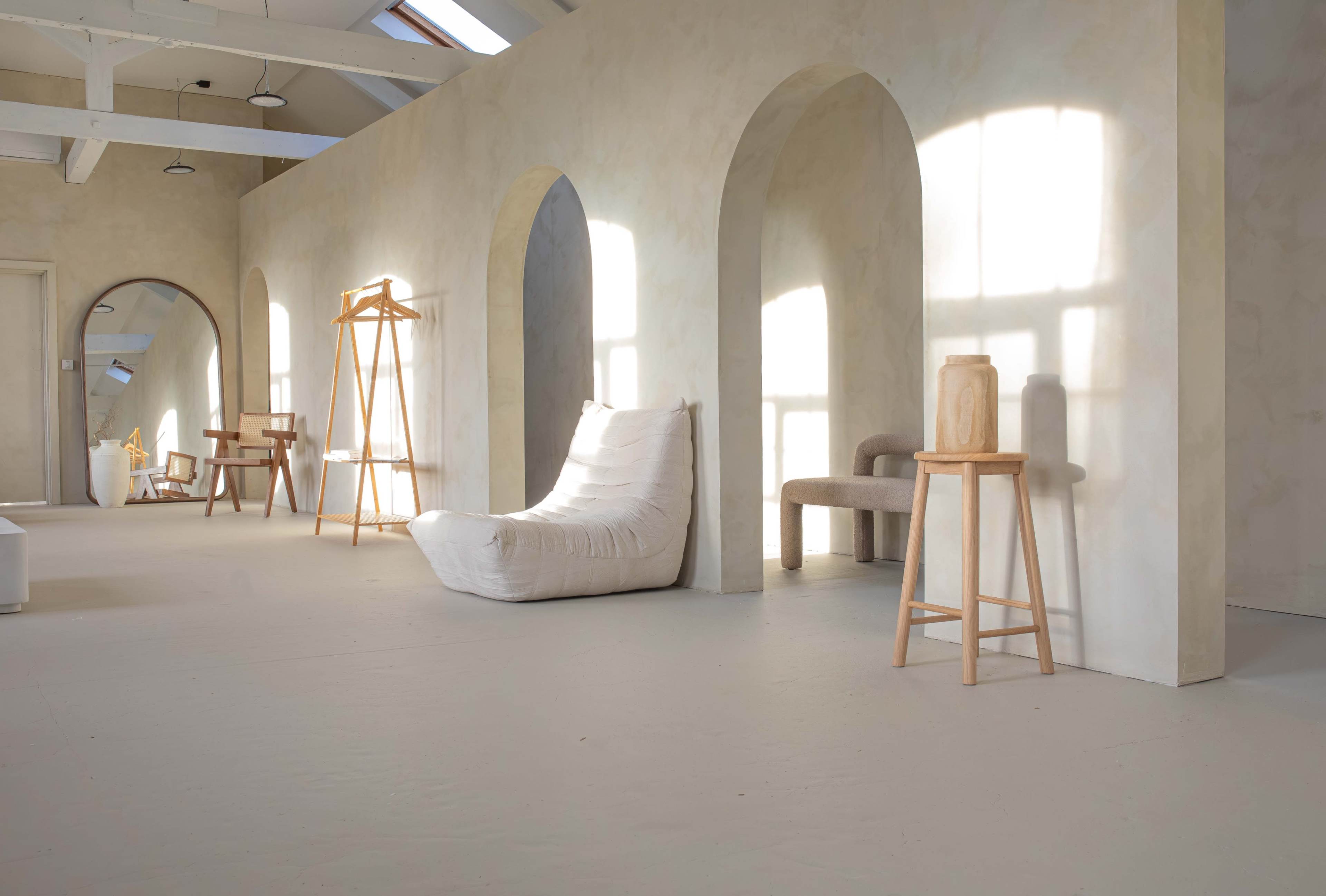 The image shows a spacious, minimalist room featuring arched openings with various wooden furniture pieces, including a chair, a coat rack, and a stool, all set against light-colored walls.