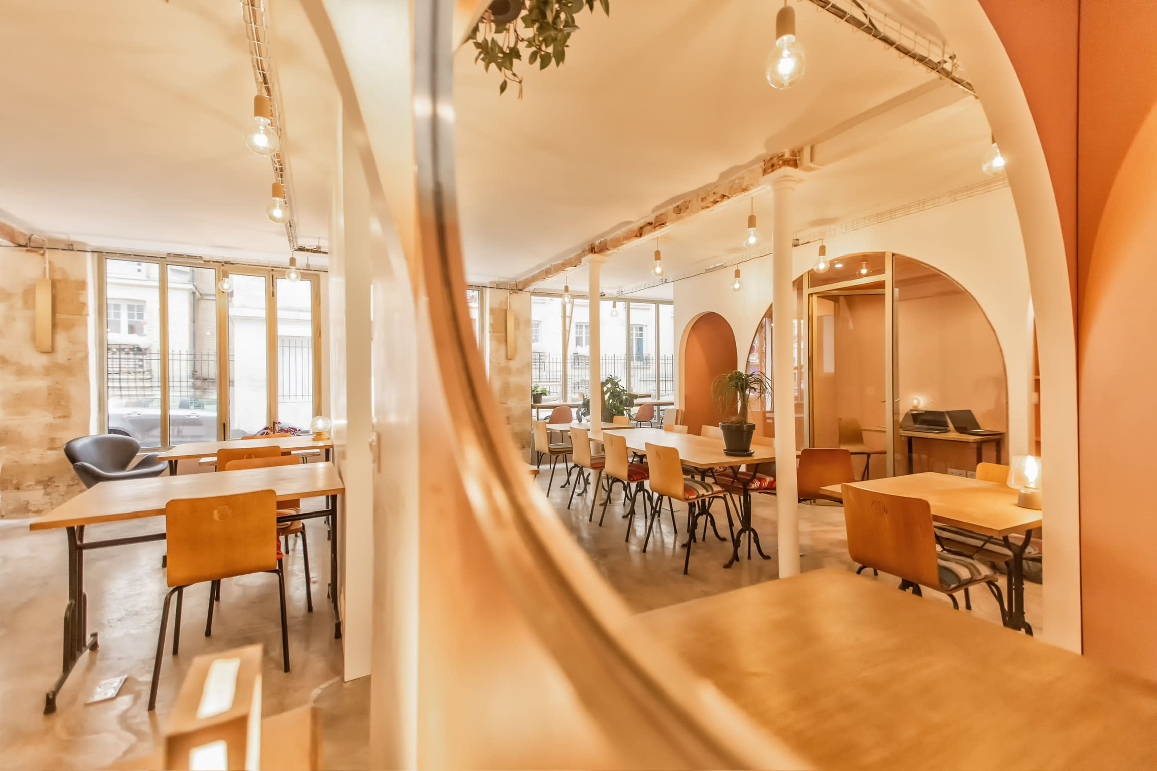 The image shows a modern, airy café with wooden tables, chairs, and warm lighting, featuring large windows and decorative plants.