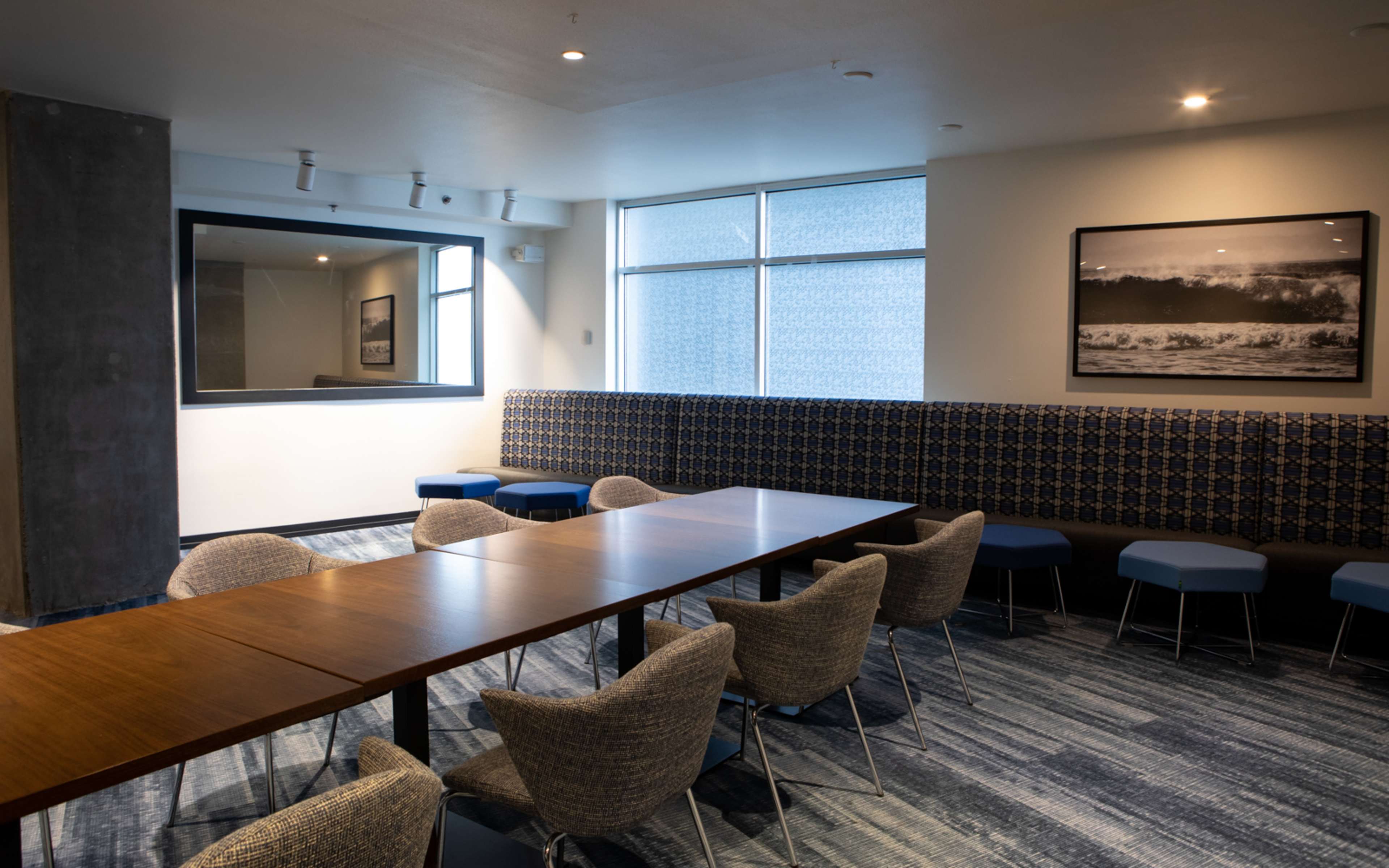 A spacious meeting room features a long wooden table surrounded by upholstered chairs, with large windows and a photograph on the wall.