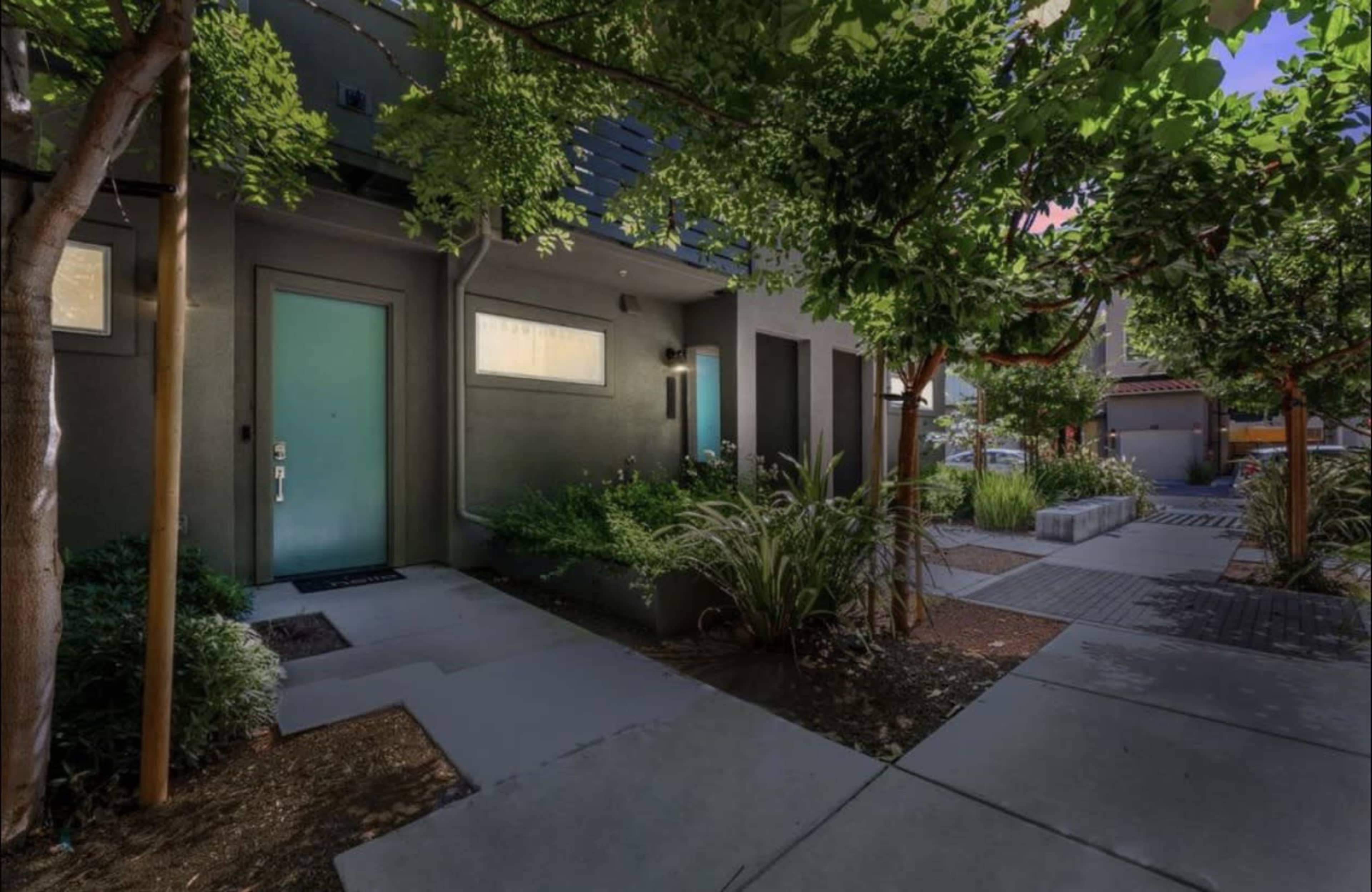 The image shows a landscaped pathway leading to modern residential doors with teal accents, surrounded by greenery and trees.