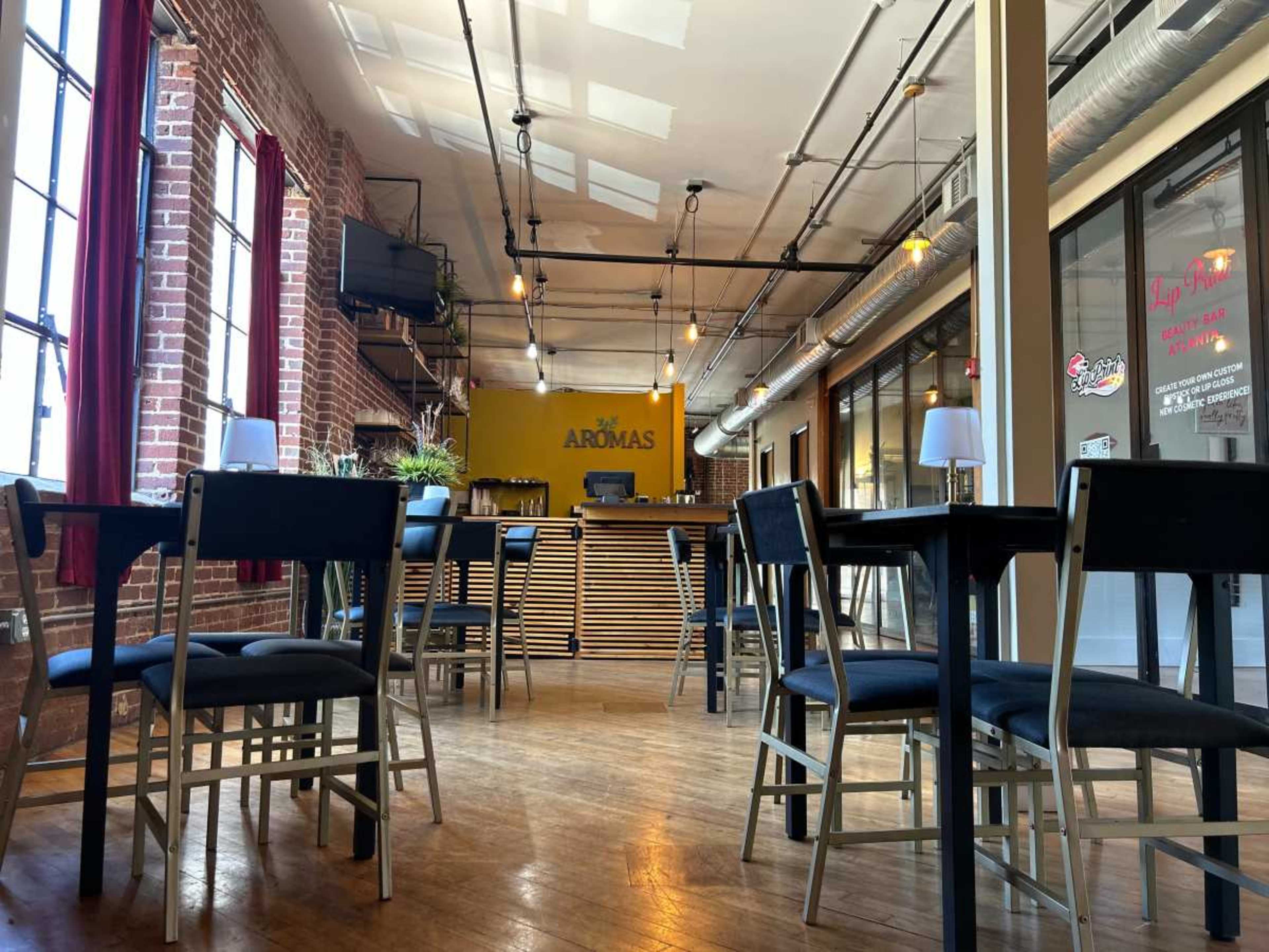 The image shows a spacious restaurant interior with wooden floors, simple tables and chairs, and a reception area featuring a sign that reads "AROMAS."