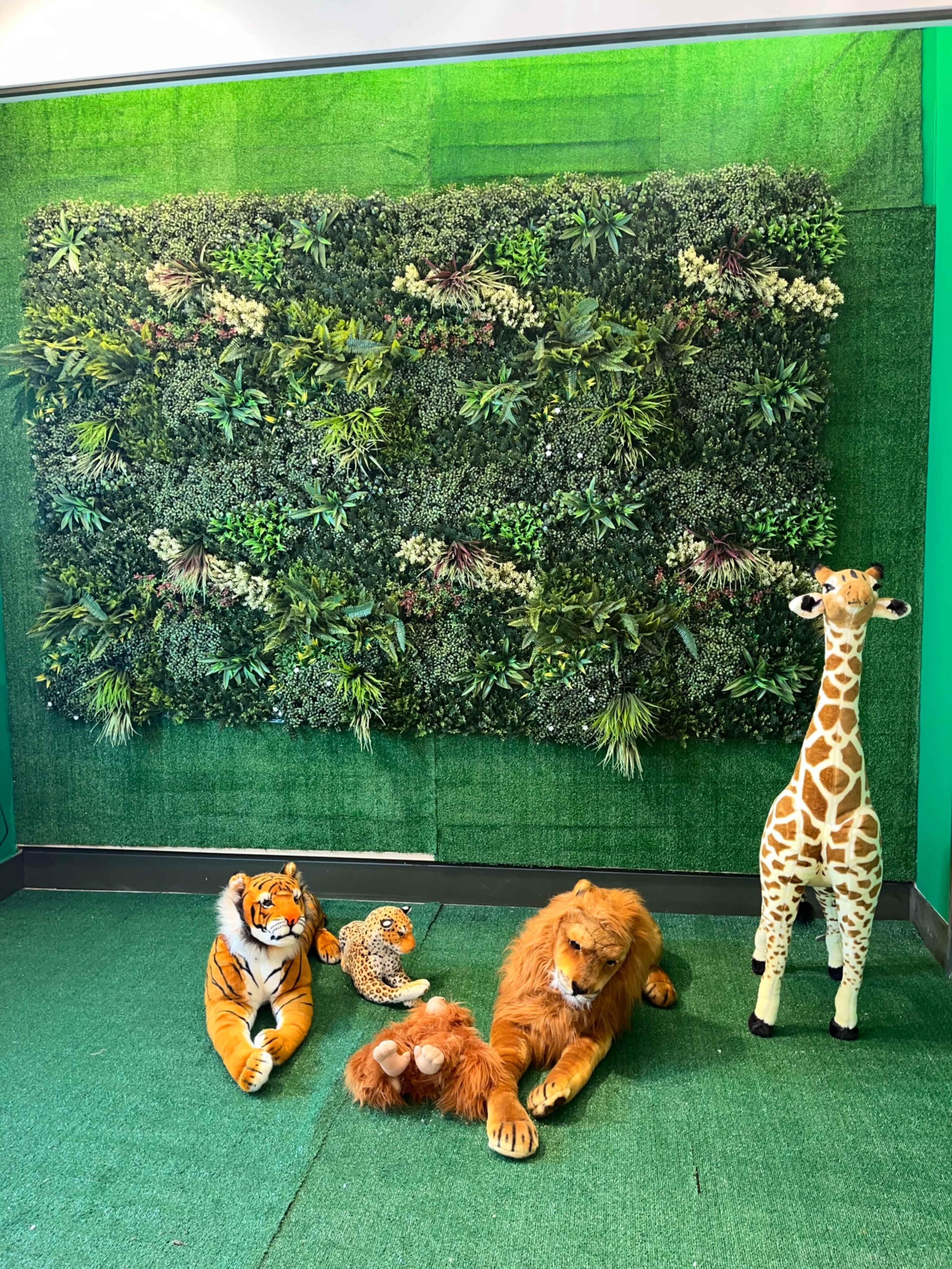 A variety of stuffed animals, including a tiger, leopard, lion, and giraffe, are arranged in front of a green leafy backdrop.