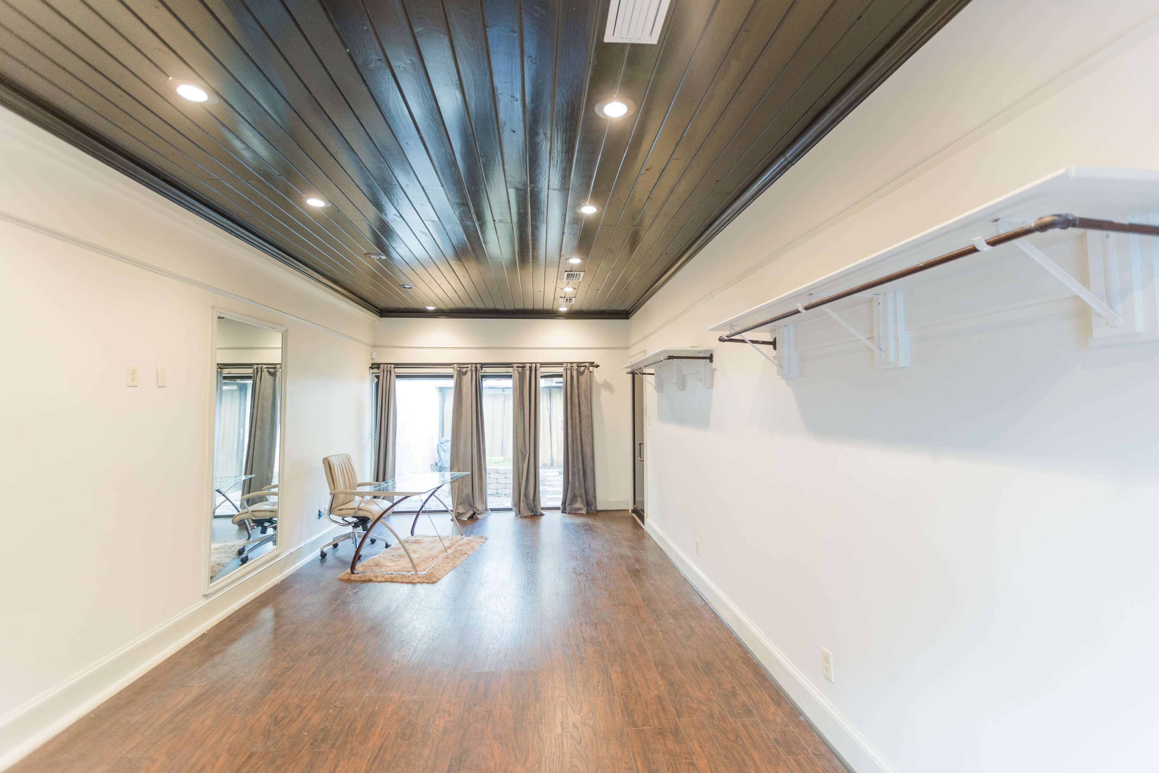 A spacious room with a dark wooden ceiling, a large mirror on one wall, and a chair positioned near a window with sheer curtains.