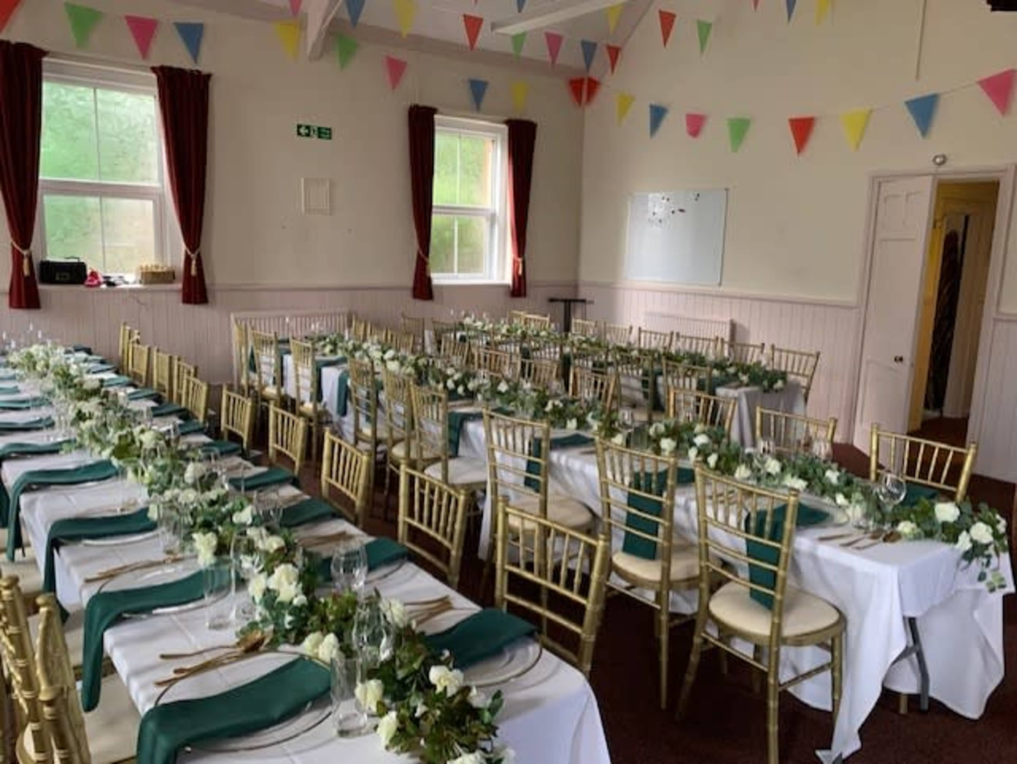 A banquet hall arranged with multiple long tables covered in white cloths, adorned with greenery and floral centerpieces, and decorated with colorful bunting overhead.