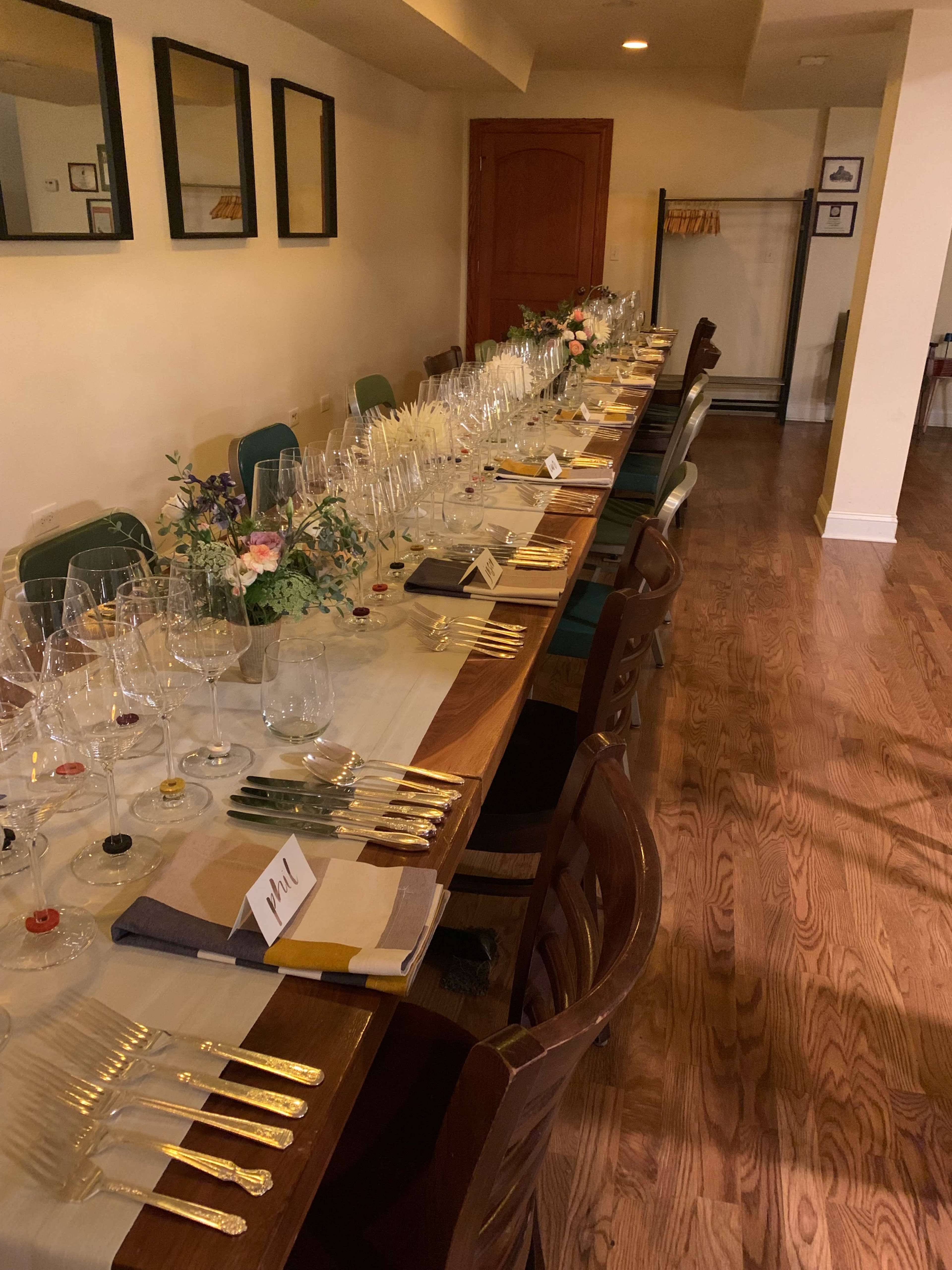 A long wooden dining table is set with glassware and silverware in a well-lit room, with framed mirrors on the wall and floral arrangements as centerpieces.