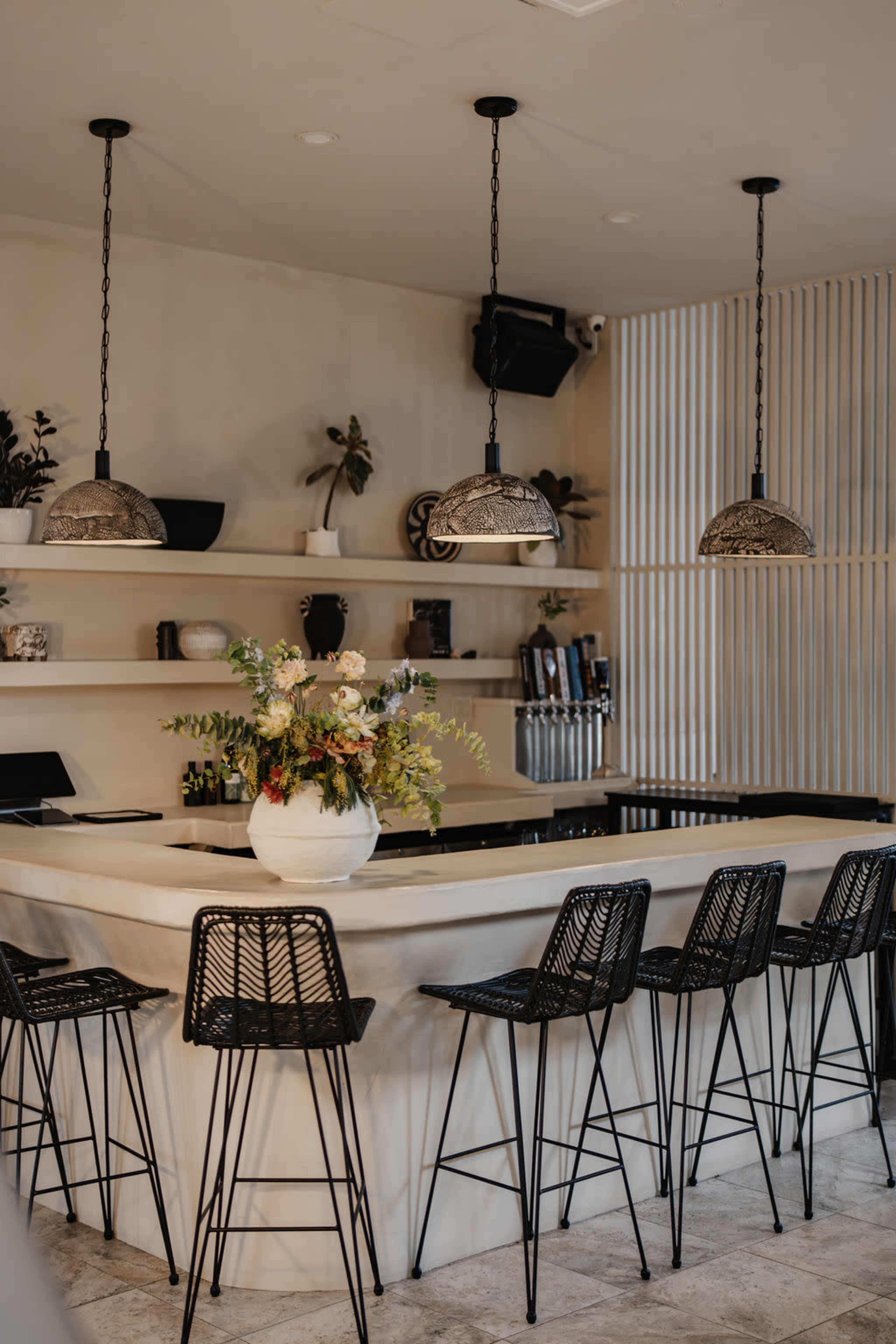 The image shows a modern bar area with a marble countertop, black wire stools, and decorative pendant lights, featuring a floral arrangement on the bar.