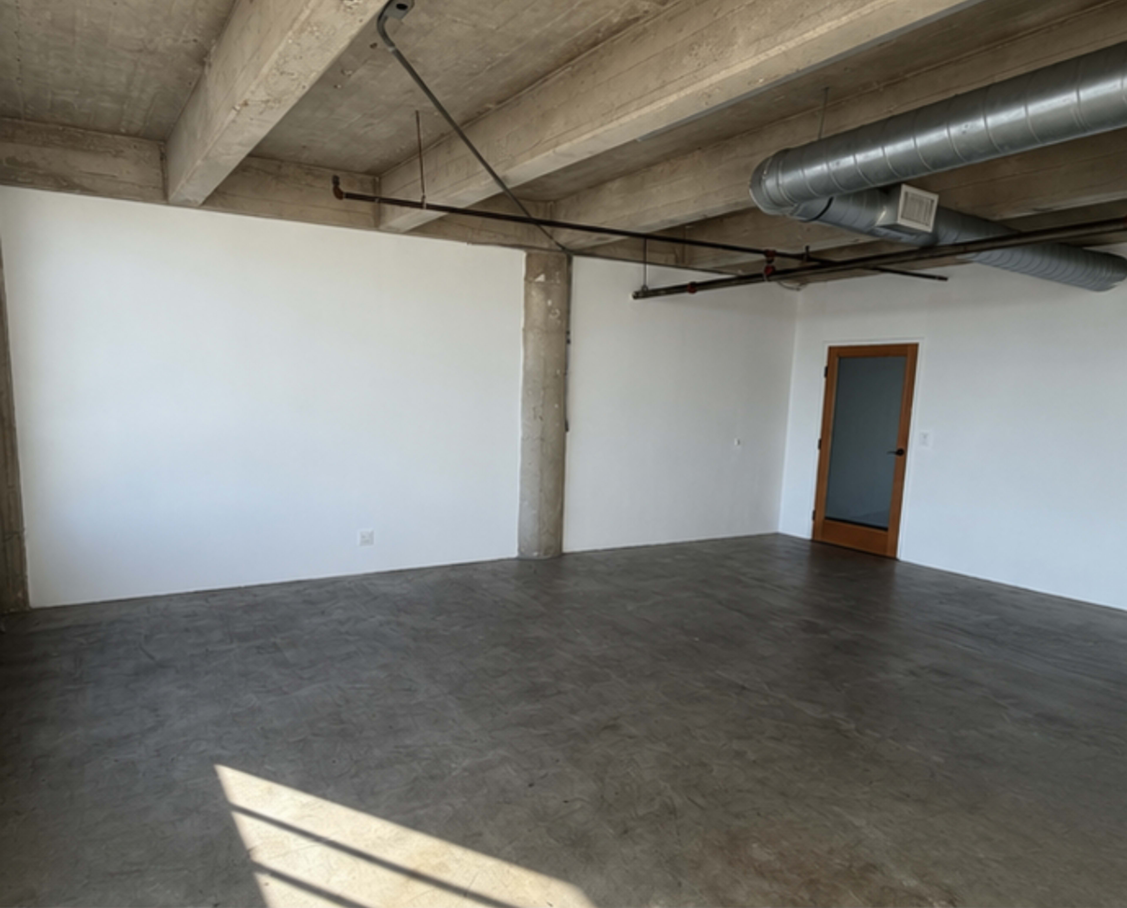 The image shows an empty room with concrete floors, exposed beams, and a glass-paned door on one side.