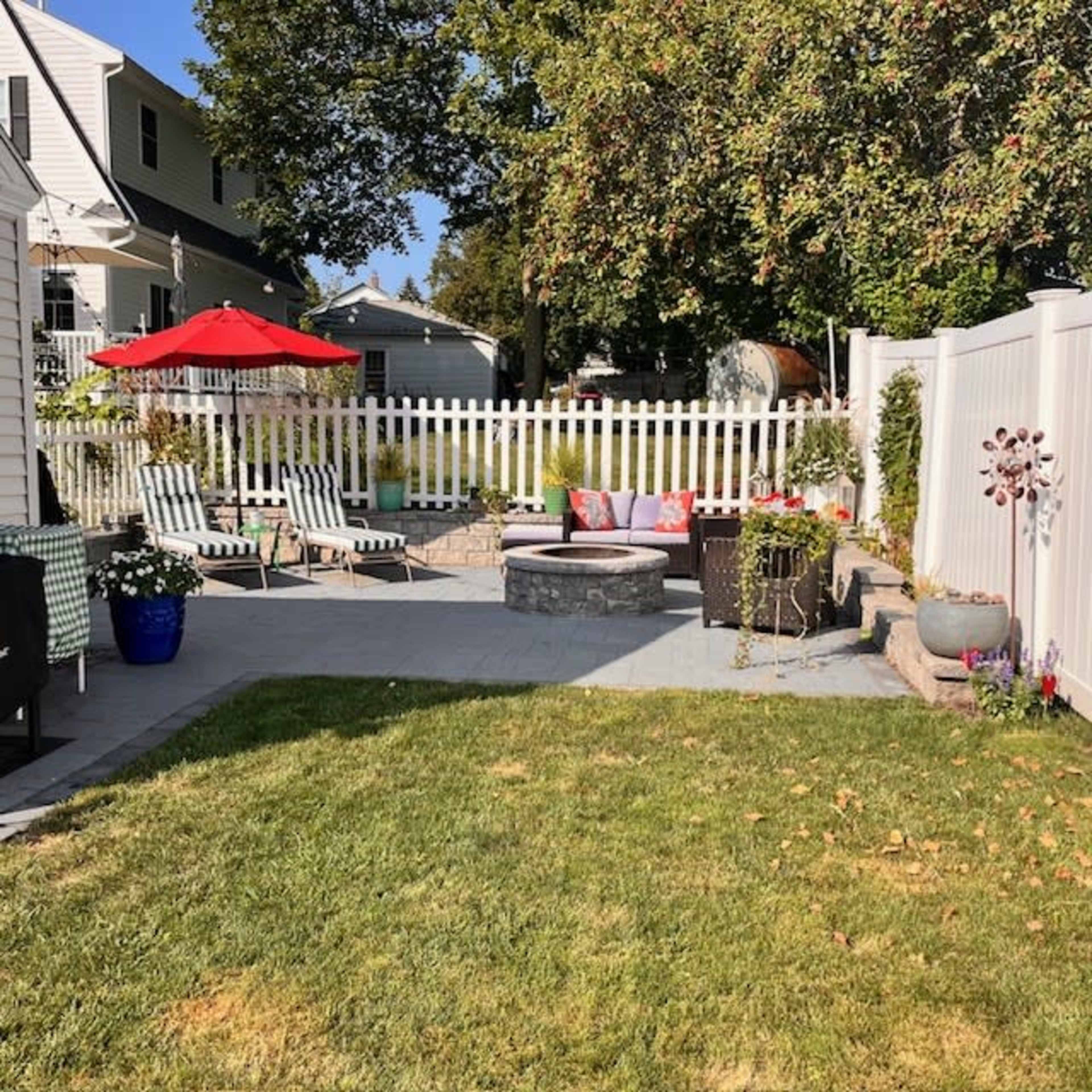 The image shows a backyard with a fire pit surrounded by seating, a well-maintained lawn, and a white picket fence, with several lounge chairs and an umbrella.