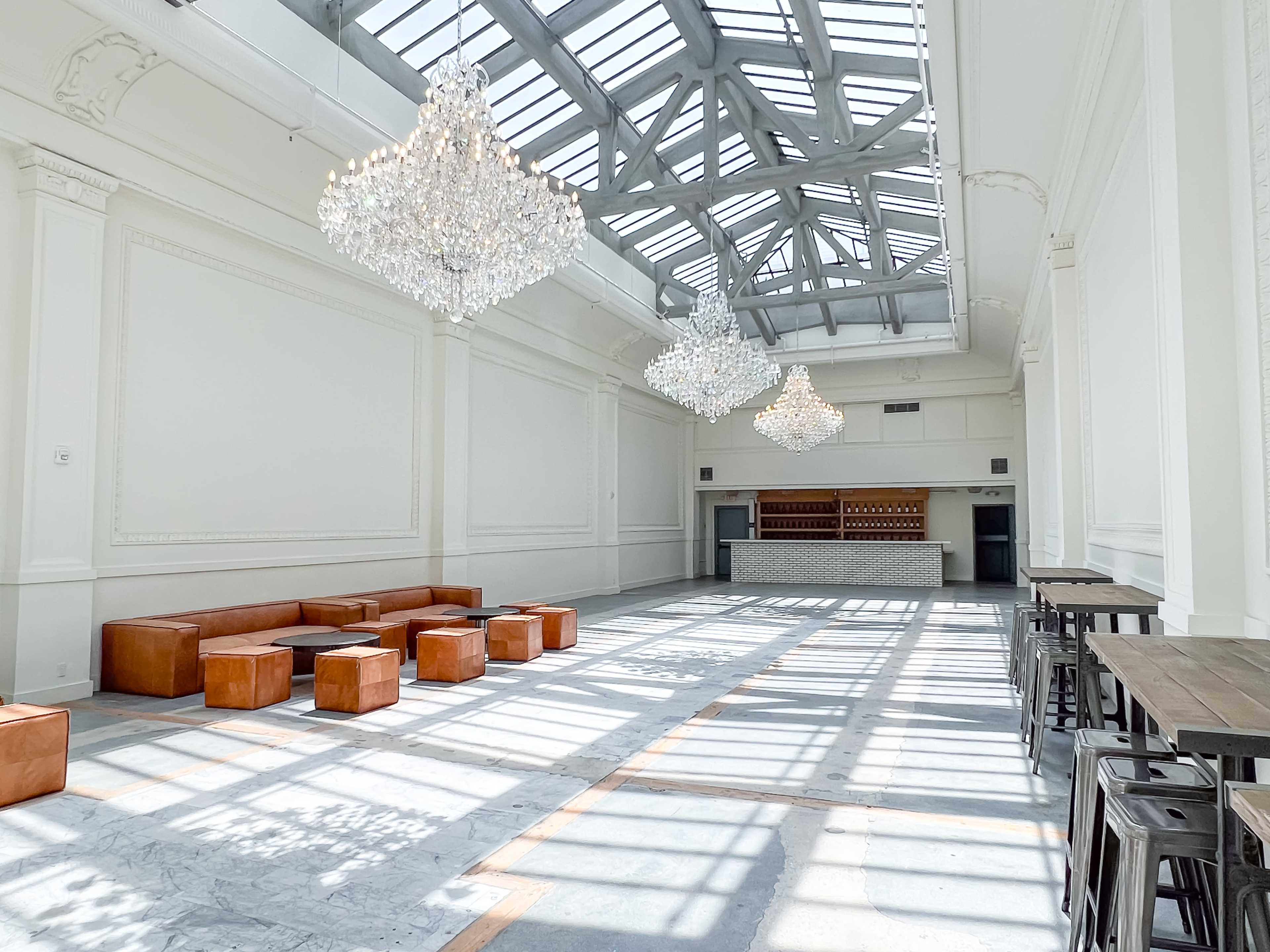 The interior features a spacious room with high ceilings, large windows, and multiple crystal chandeliers, along with wooden seating and a bar area at the back.