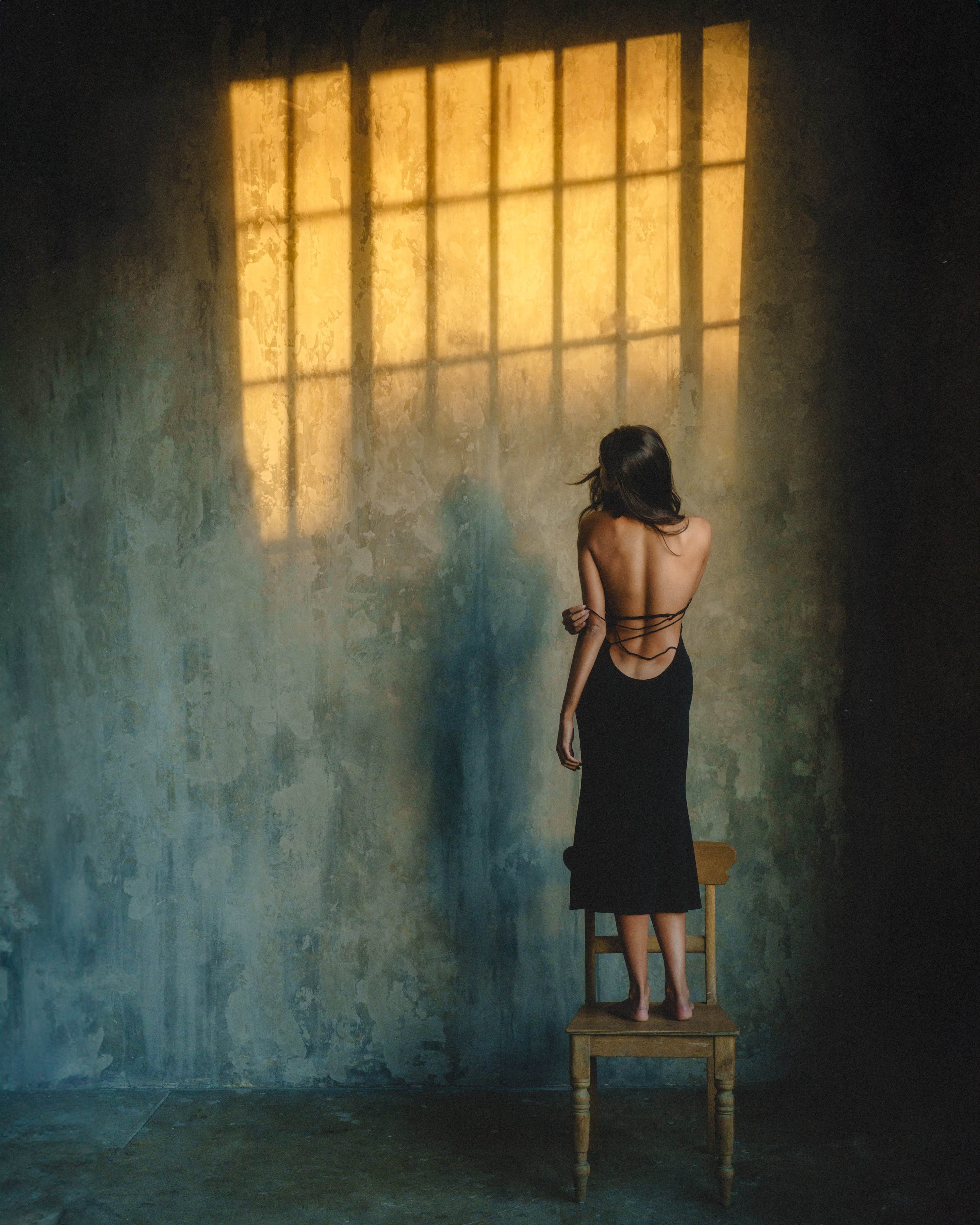 A person stands on a chair against a textured wall, with sunlight casting a grid pattern on the surface.