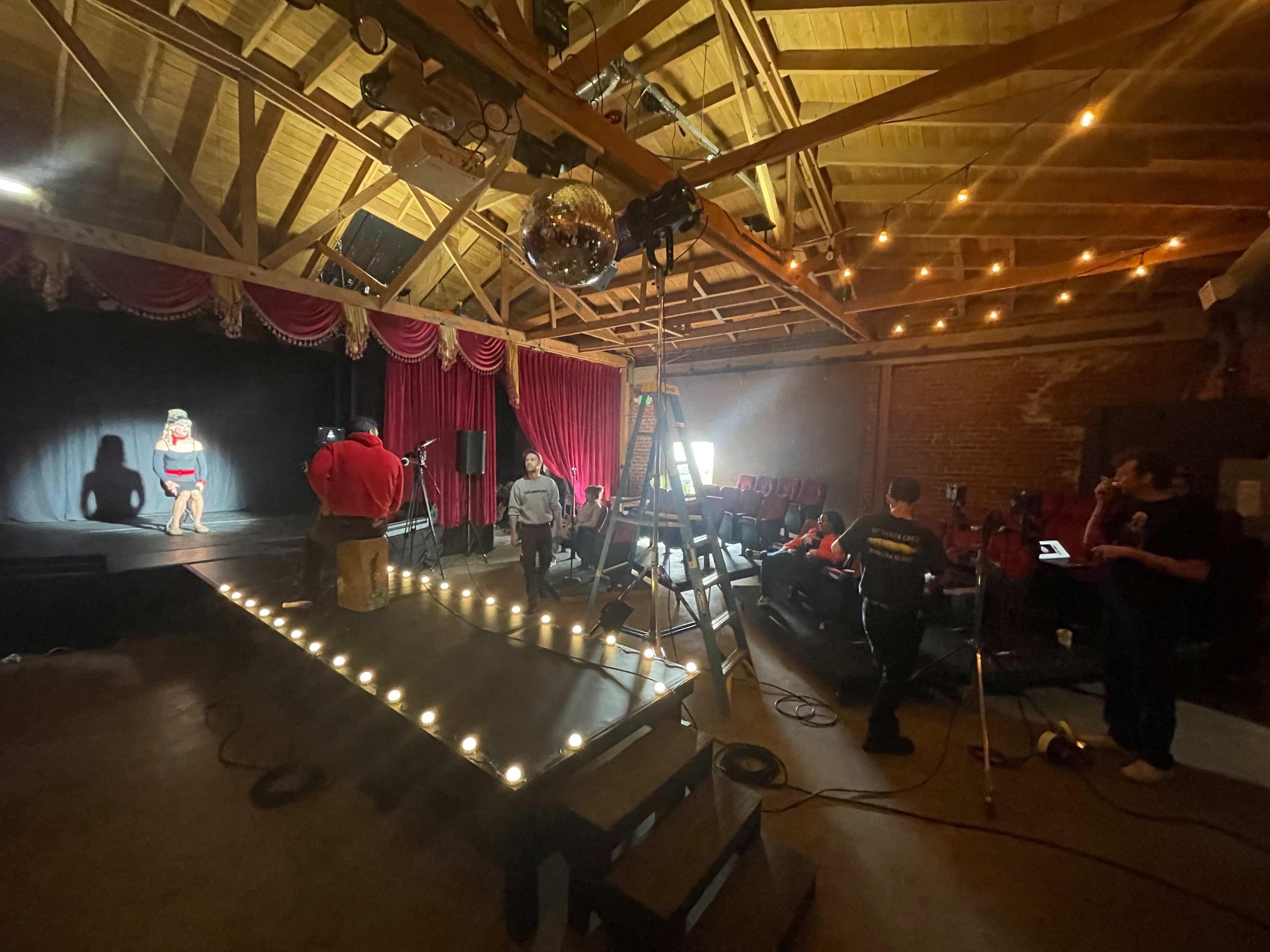 A performer on stage rehearses under lights in a rustic venue with a disco ball, while a crew sets up equipment and an audience observes.