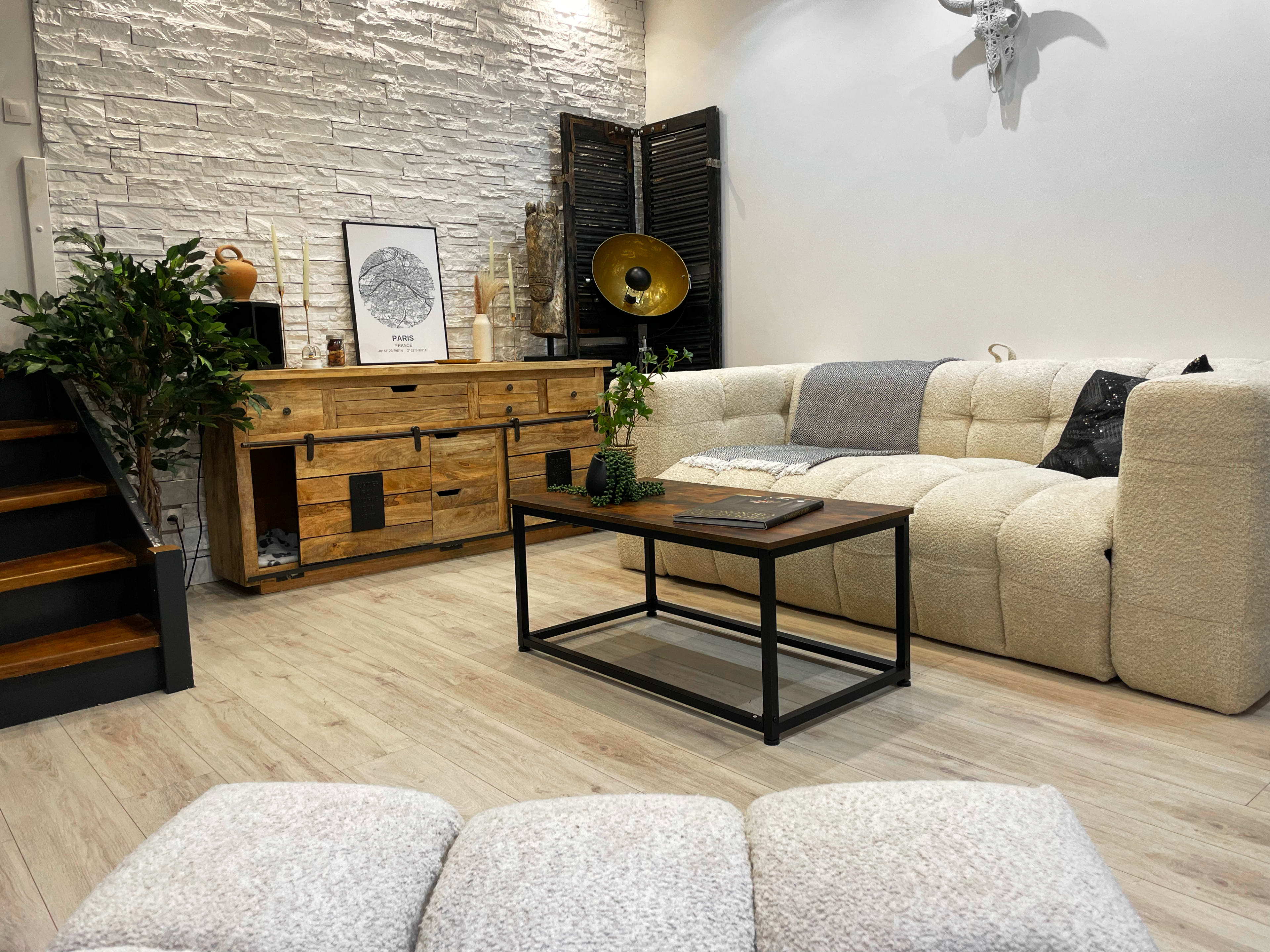 The image shows a modern living room featuring a beige upholstered sofa, a wooden coffee table, an assembly of decorative items on a sideboard, and a staircase leading to another level.