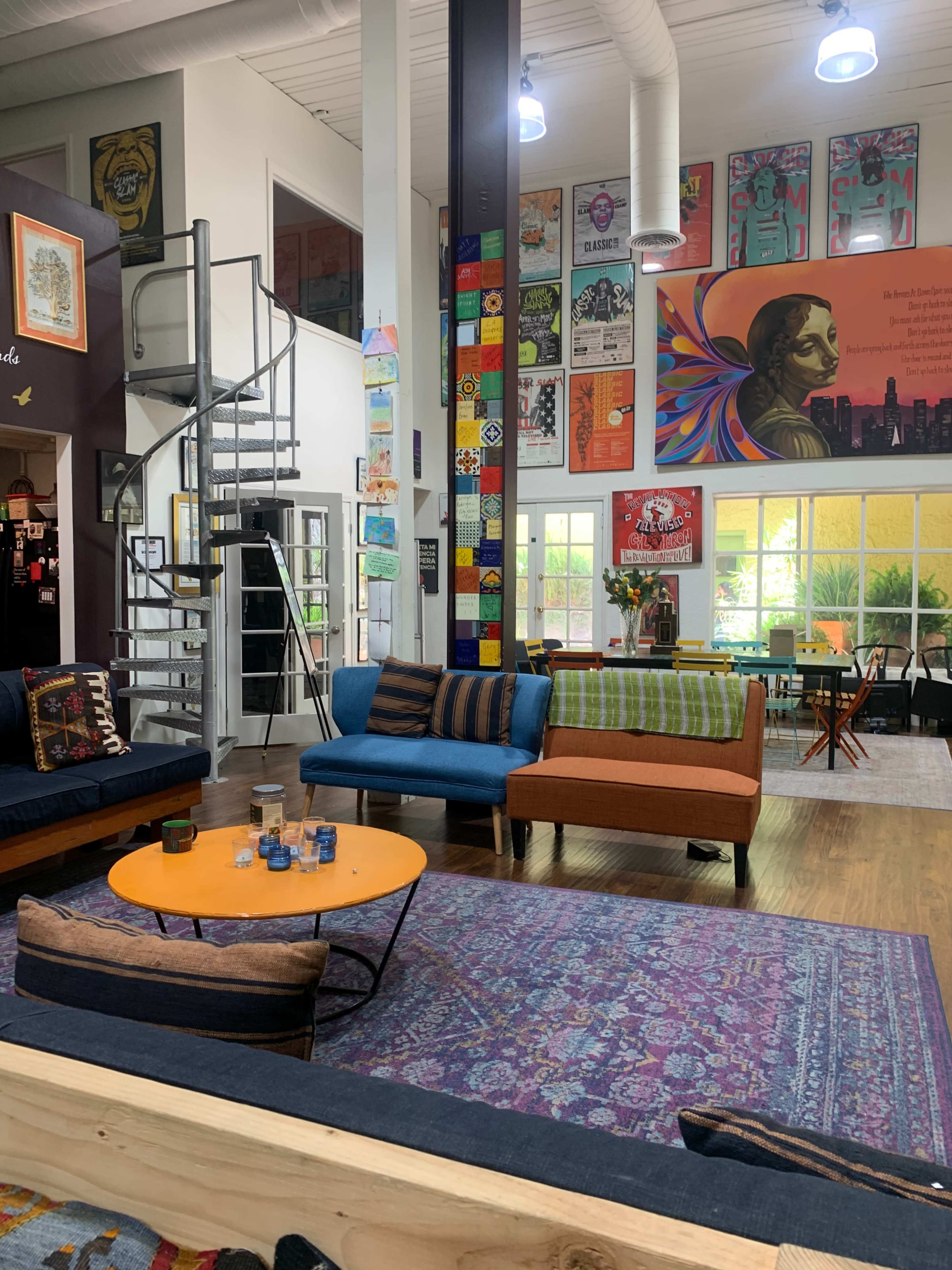 The image depicts a vibrant, open lounge area with colorful furniture, a spiral staircase, and walls adorned with various artistic posters.