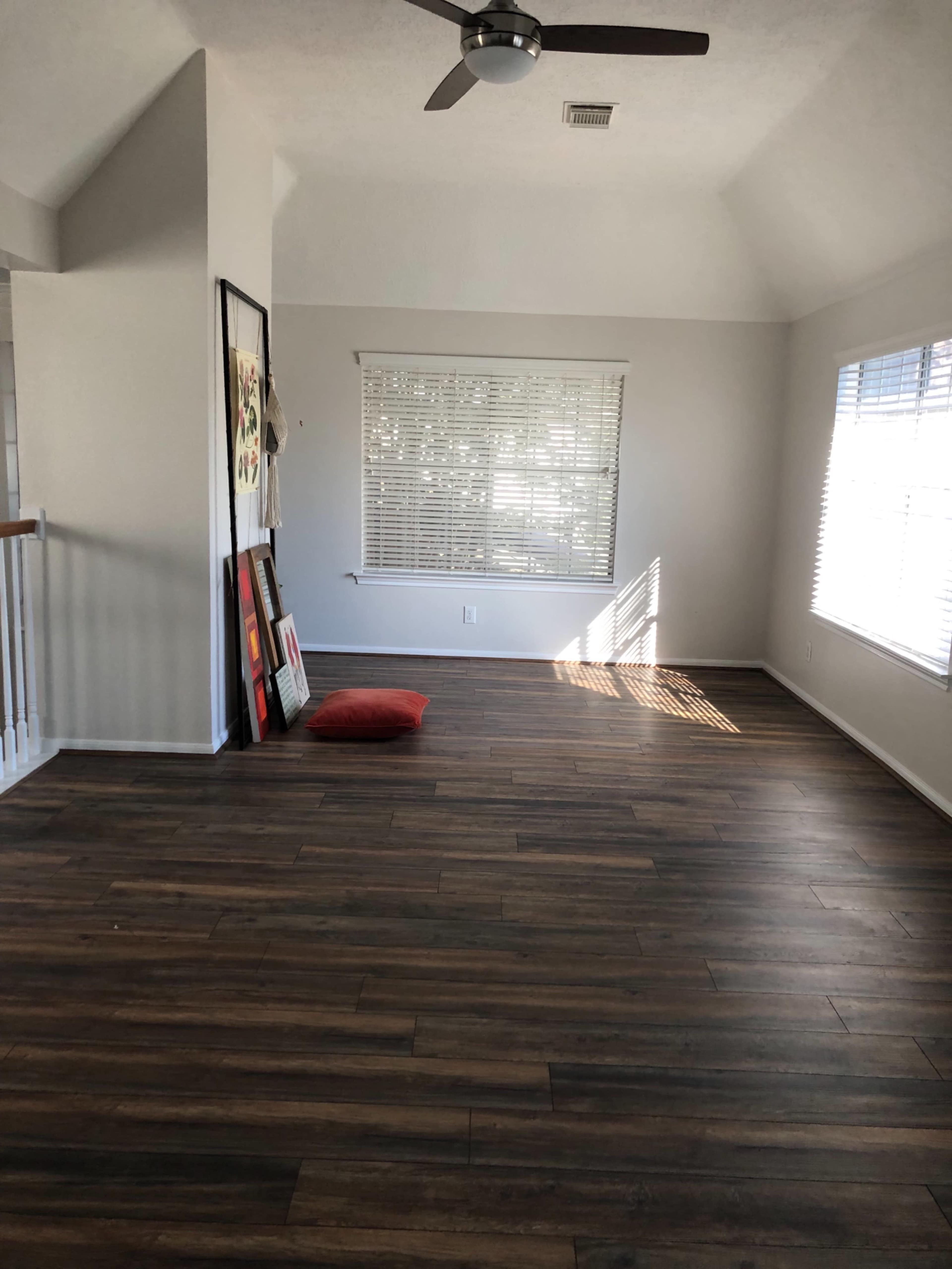 An empty room with wood-like floors, a ceiling fan, large windows with blinds, and a red cushion on the floor near a wall with framed artwork.
