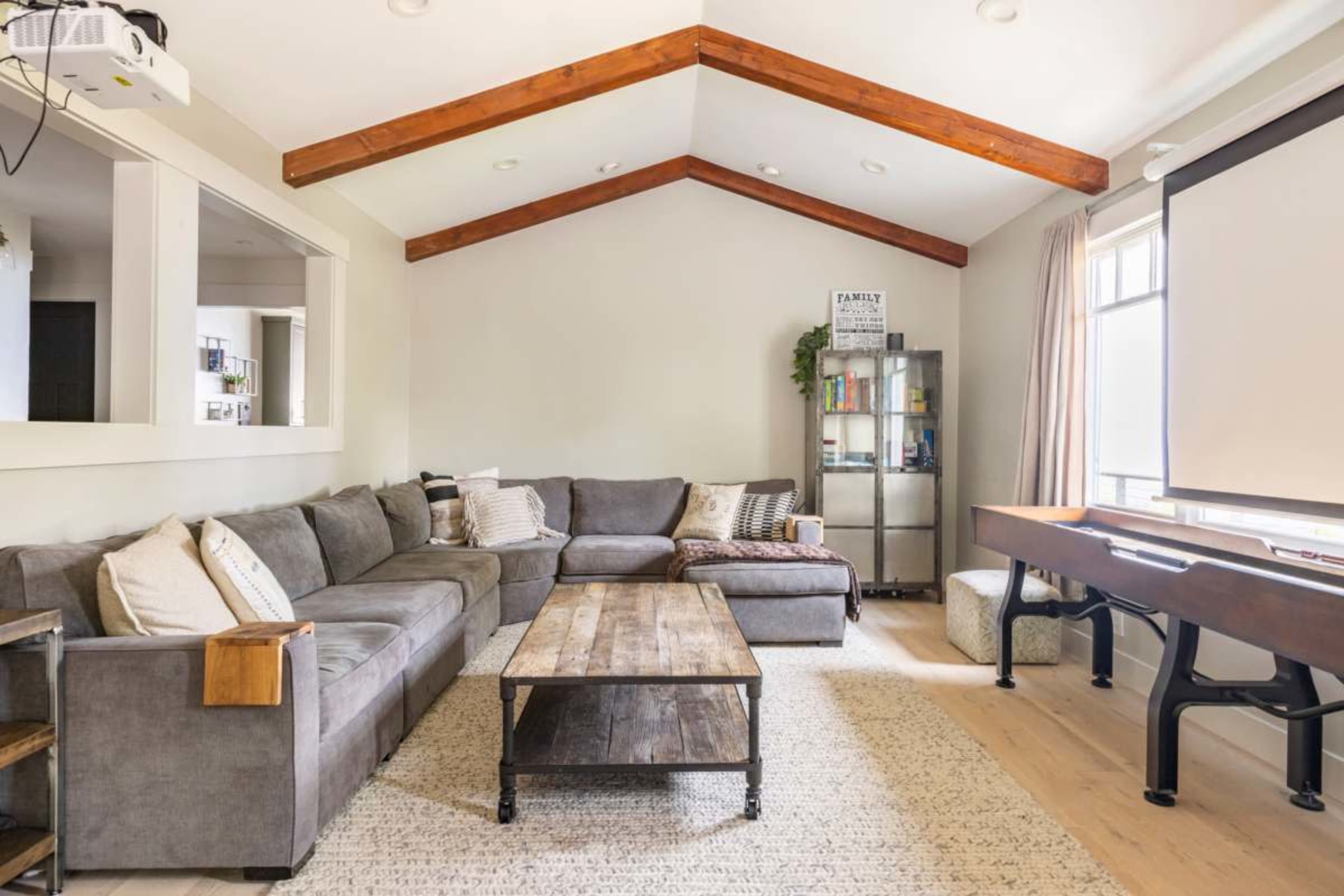 A spacious living room features a large sectional sofa, a wooden coffee table, and a projector screen on one wall with exposed wooden beams overhead.