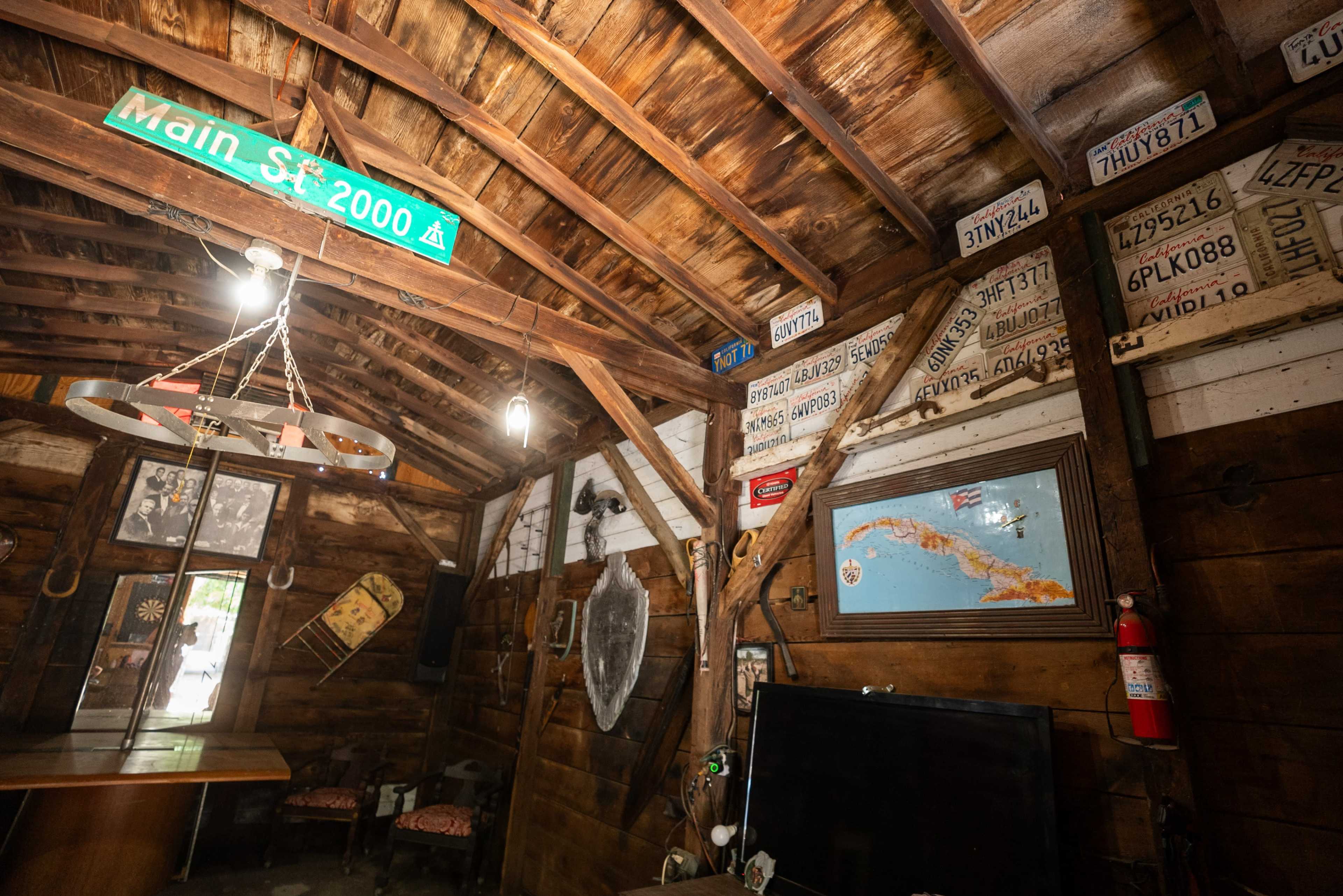 The interior of a rustic cabin features wooden beams, a table, a television, and various vintage license plates on the walls.