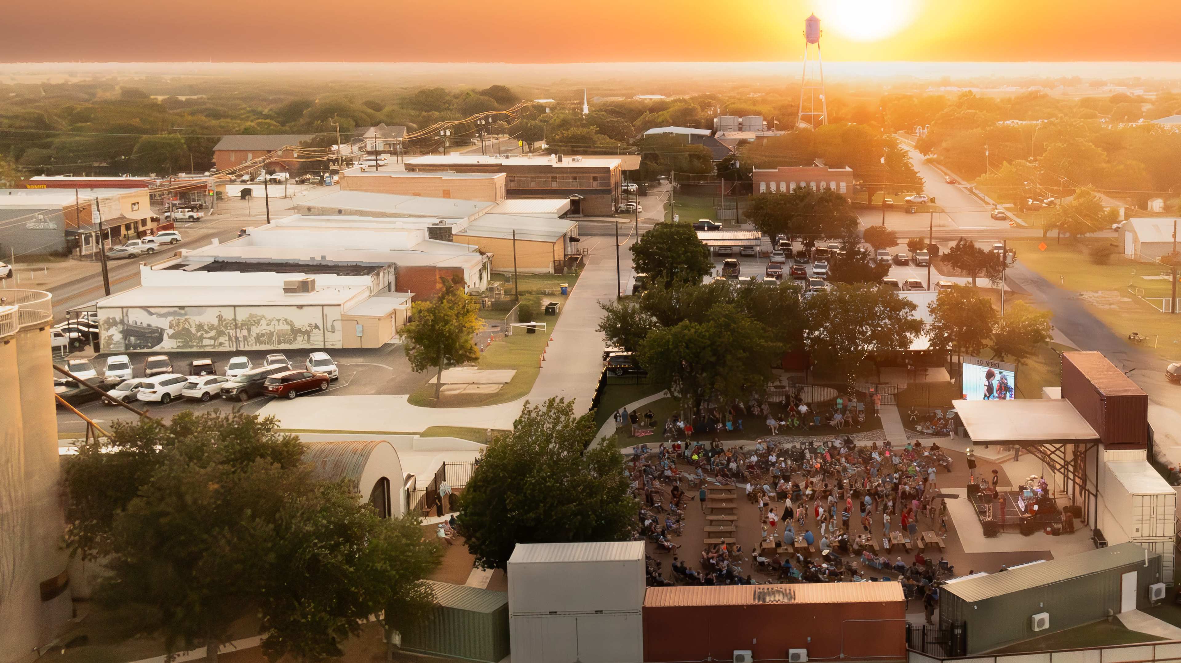 The image shows a large outdoor gathering at sunset in a town center, with people seated in a park area facing a screen amidst surrounding buildings.