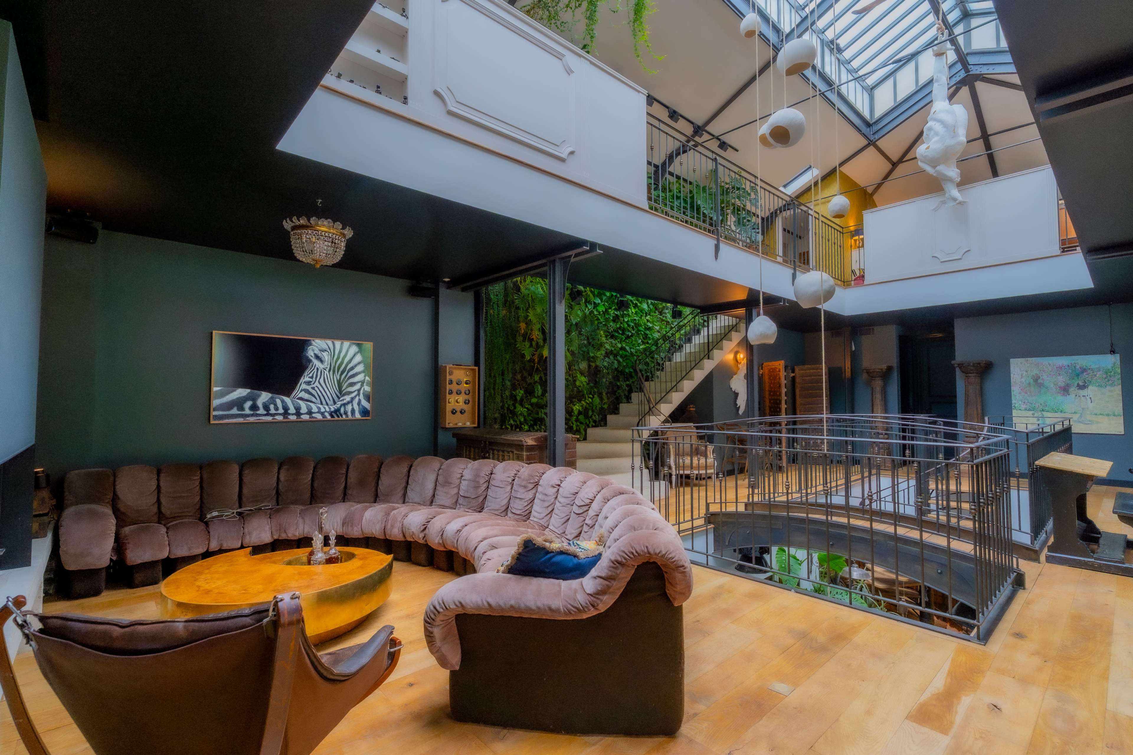 An open living space features a large curved sofa, a round wooden table, and a staircase leading to a second level, all surrounded by lush greenery and modern decor.