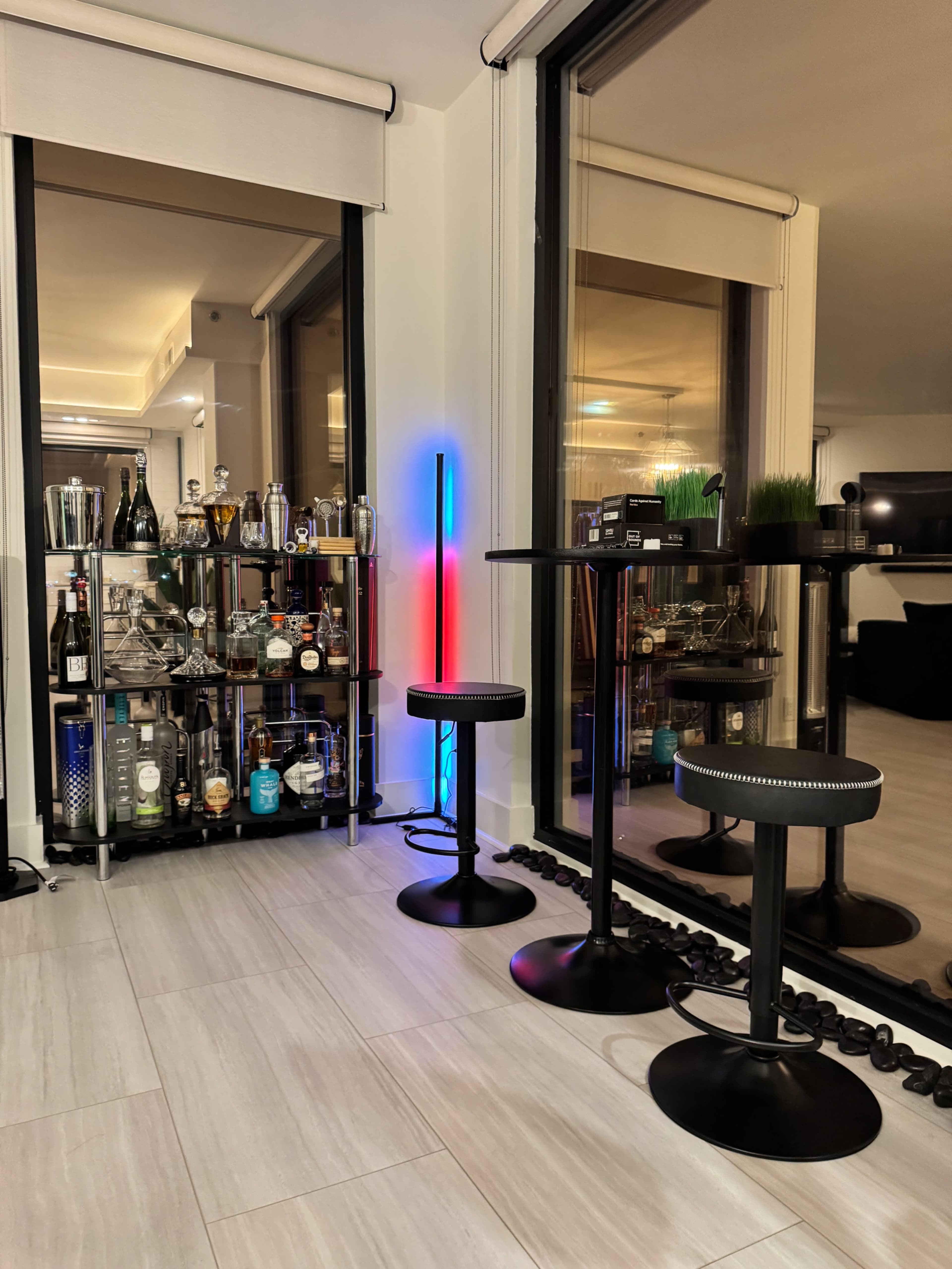 A home bar area features a shelf filled with various bottles, a colorful LED light, and two black stools beside a small round table.
