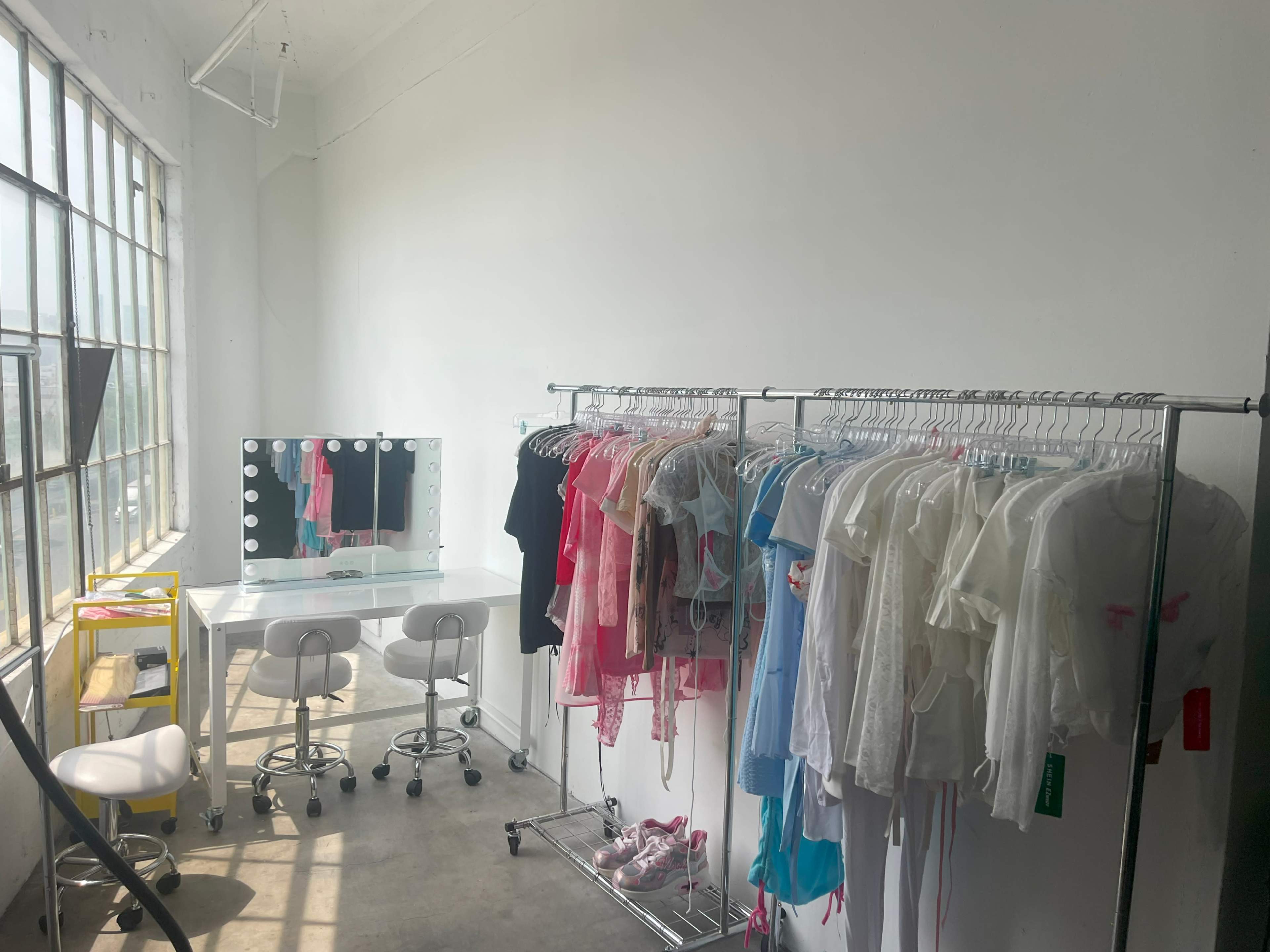 A well-lit room with large windows, featuring clothing racks filled with various garments and a vanity table with mirrors and seating.