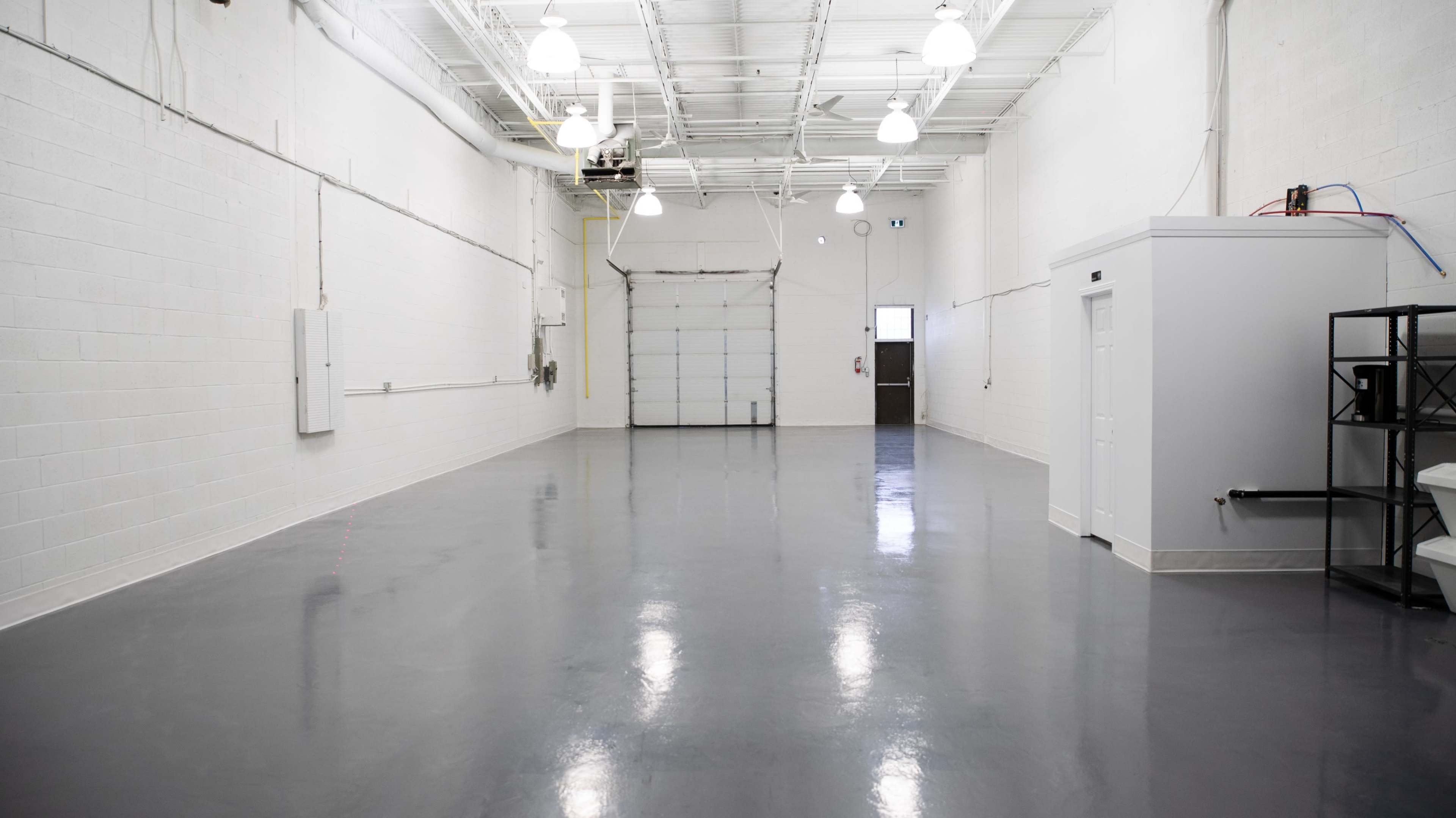 A large, empty industrial space with glossy gray flooring, white walls, and a roll-up garage door at one end.