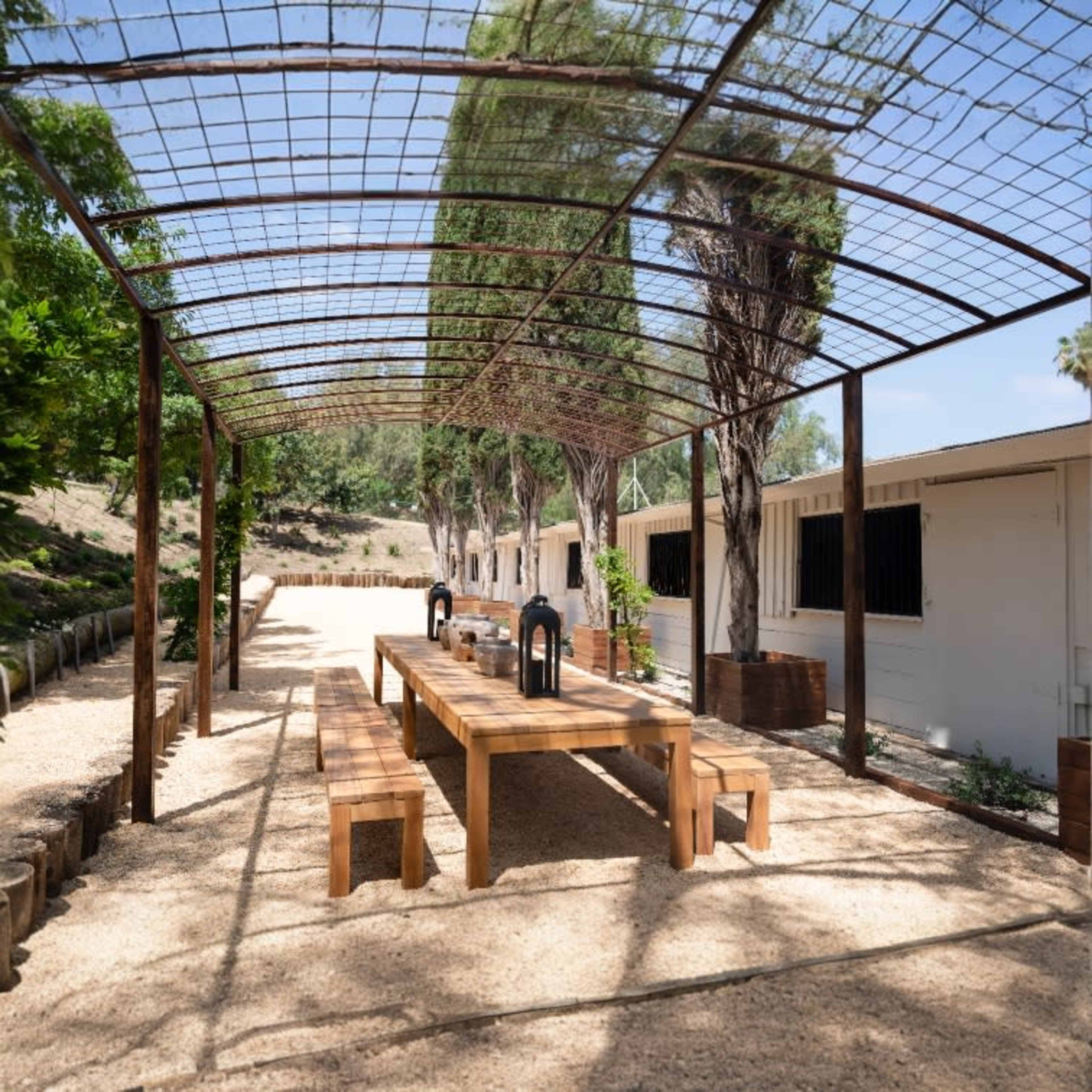 A long wooden table is set under a trellis in a gravel area surrounded by trees and a building in the background.