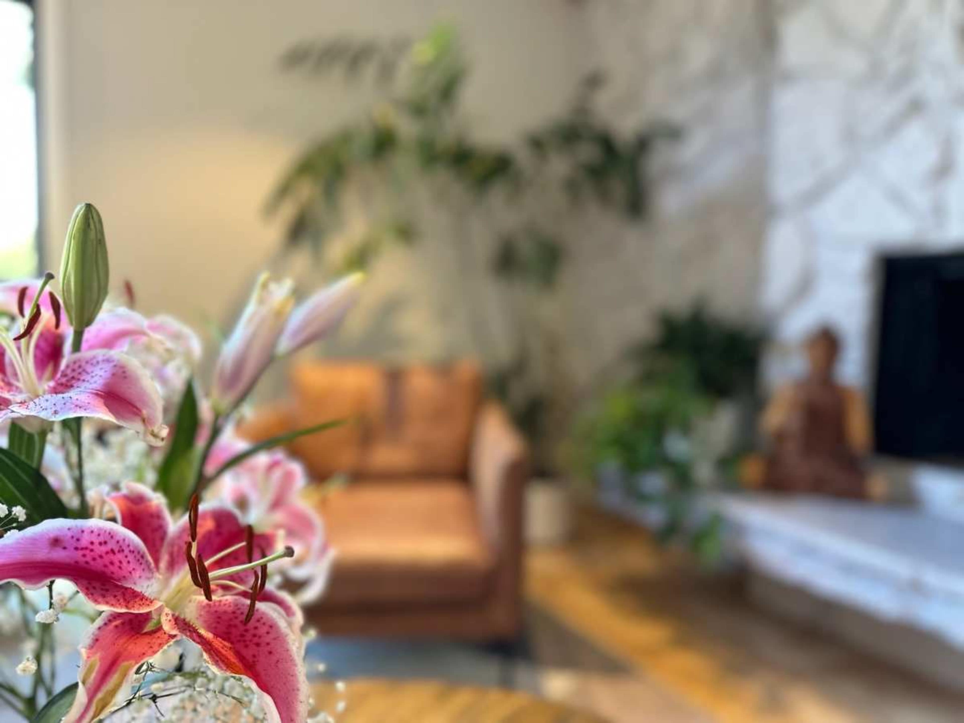 A blurred bouquet of pink lilies is in the foreground, with a brown sofa and potted plants visible in a bright, modern living room in the background.