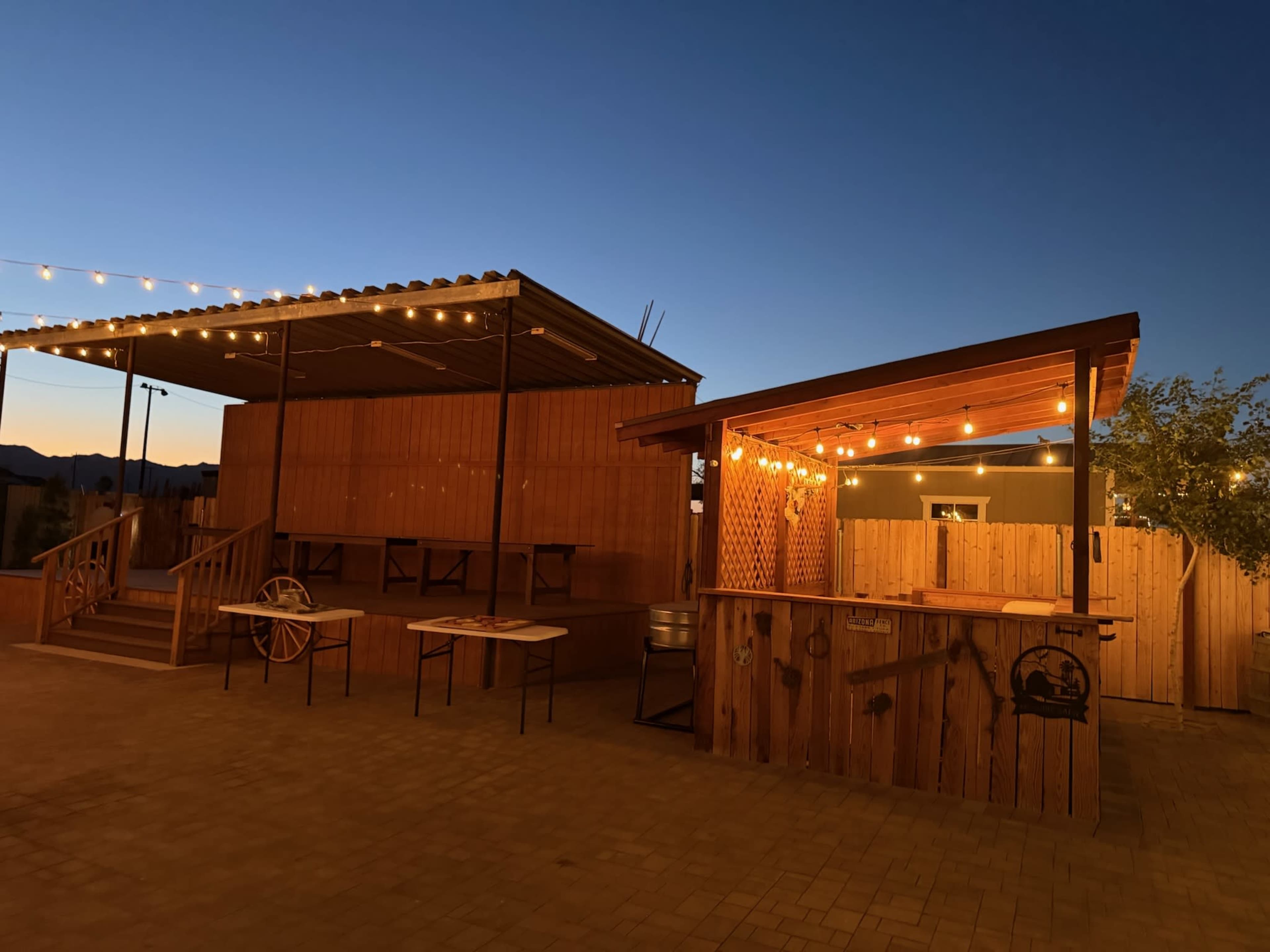 The image shows a wooden outdoor space with a bar area and a raised platform, illuminated by string lights against a twilight sky.