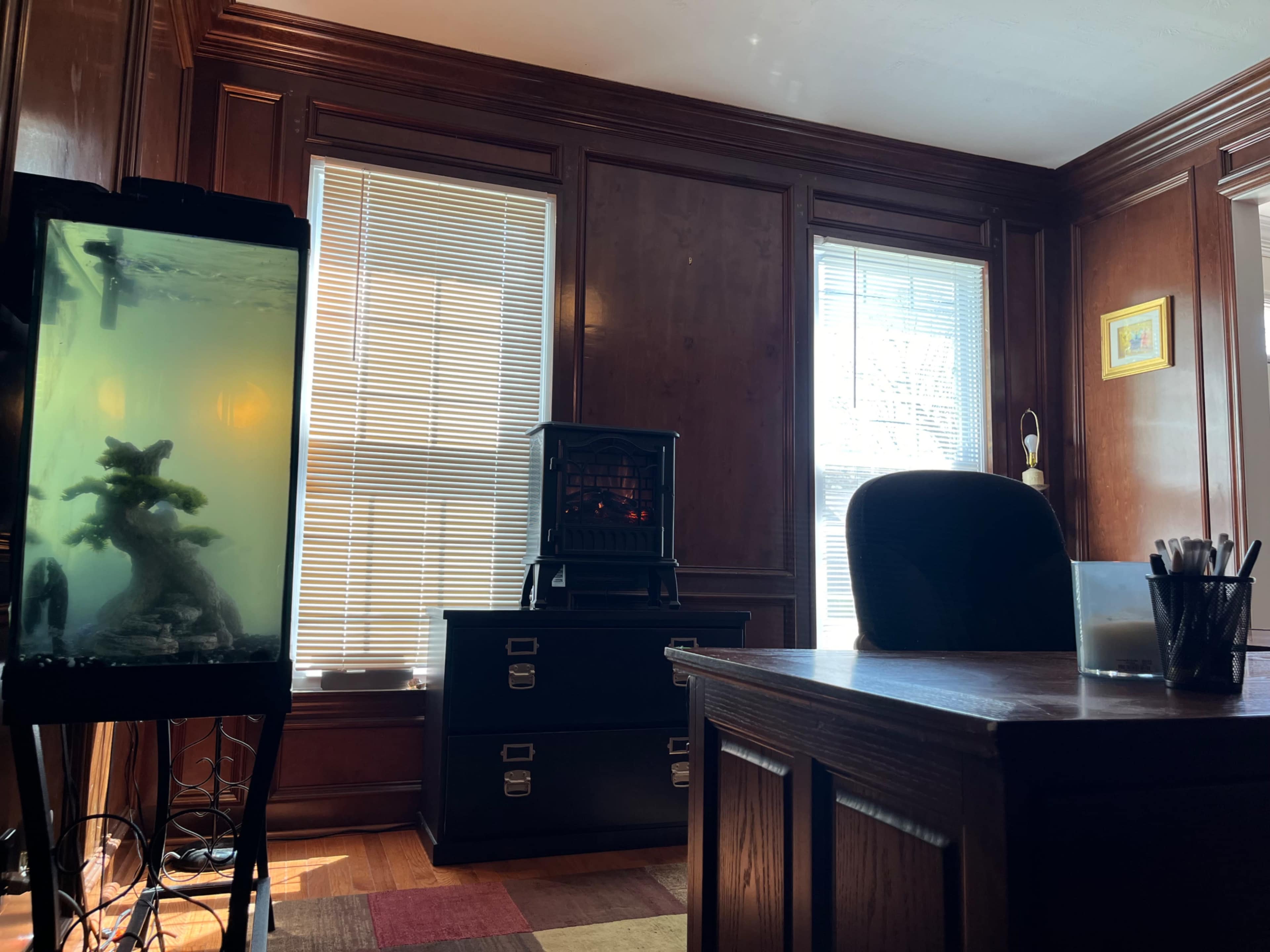 A wooden office with a fish tank on the left, a black cabinet in the center featuring a decorative heater, and a desk on the right.