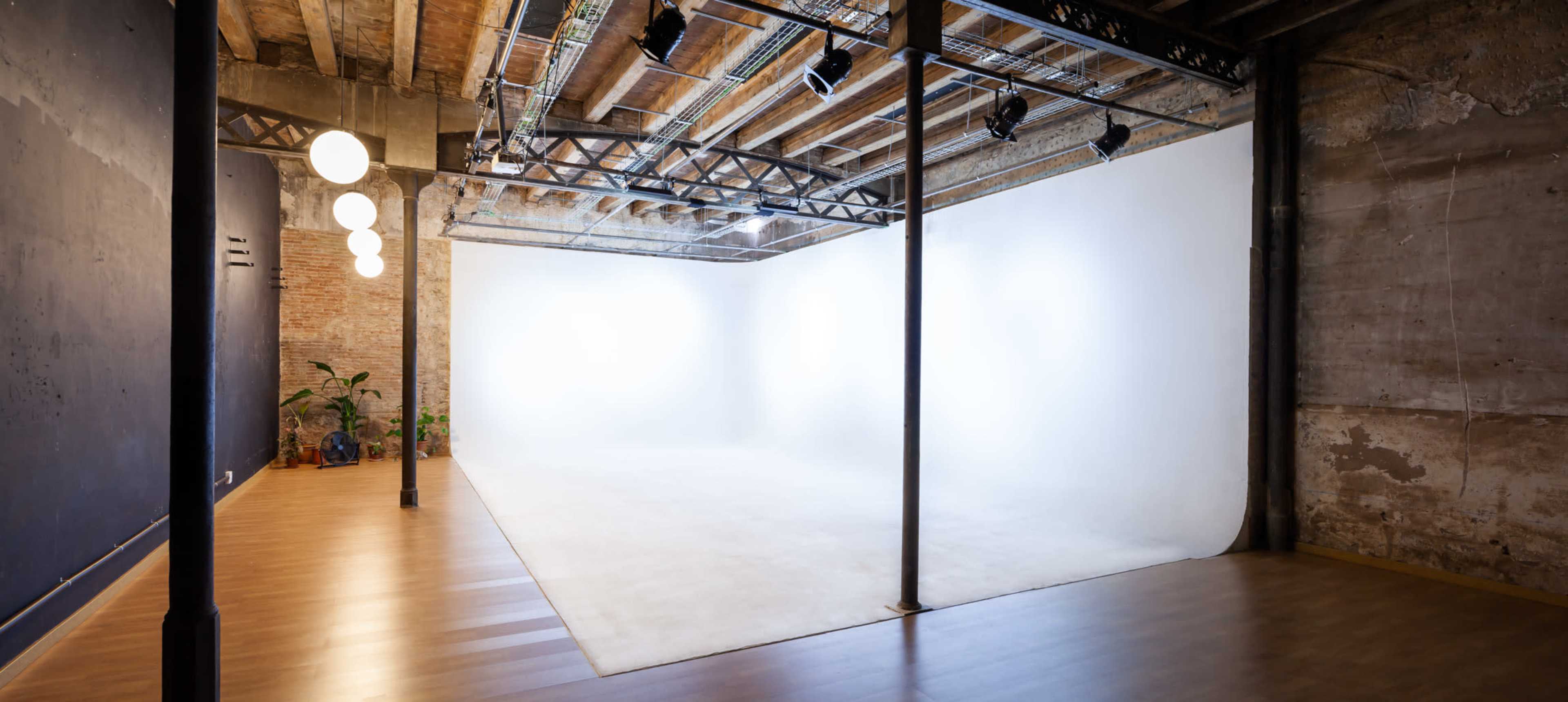 An empty studio space features a white backdrop, wooden flooring, and overhead lighting fixtures.