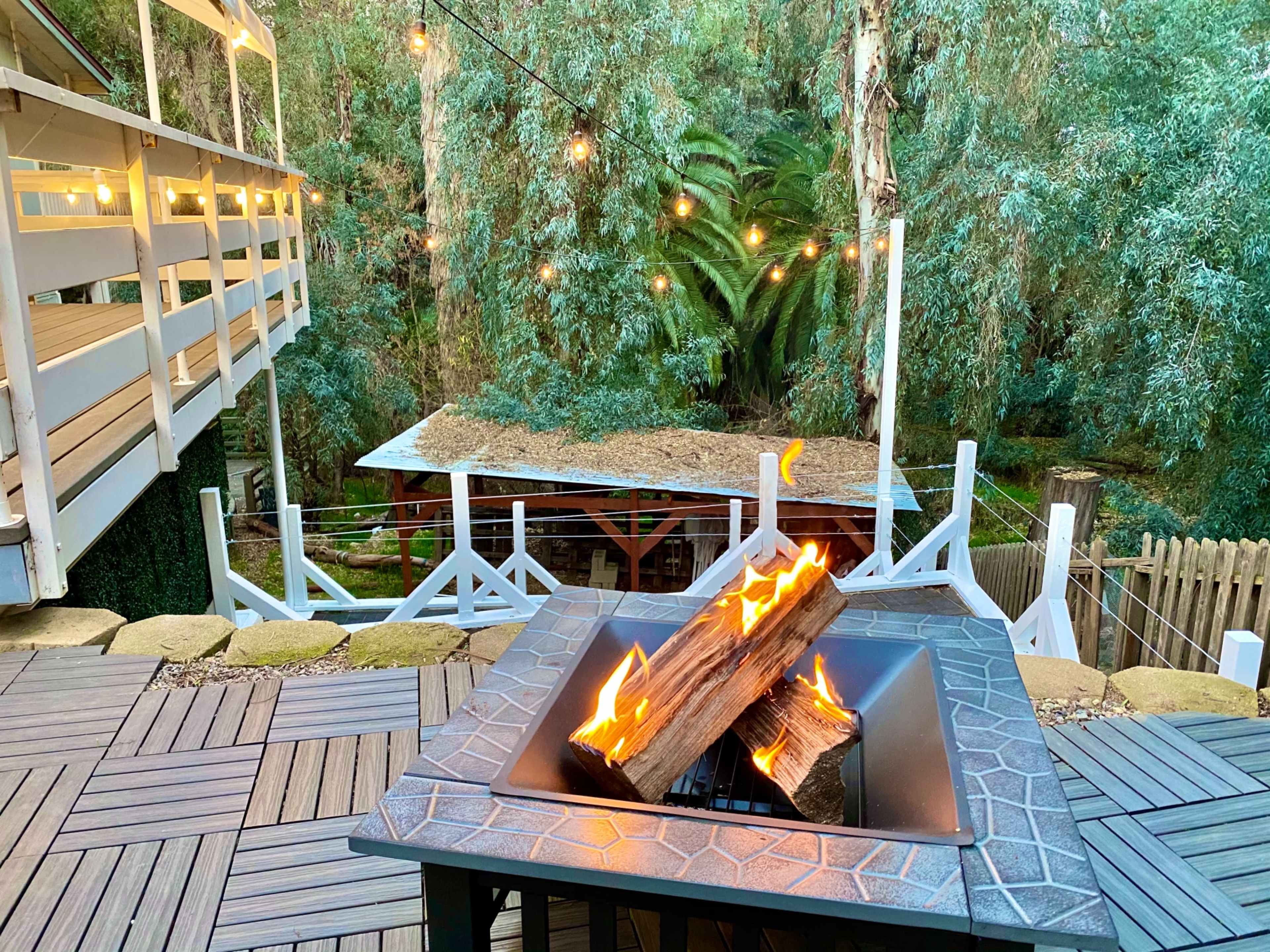 A fire pit with burning logs sits on a wooden deck surrounded by trees and string lights in the background.