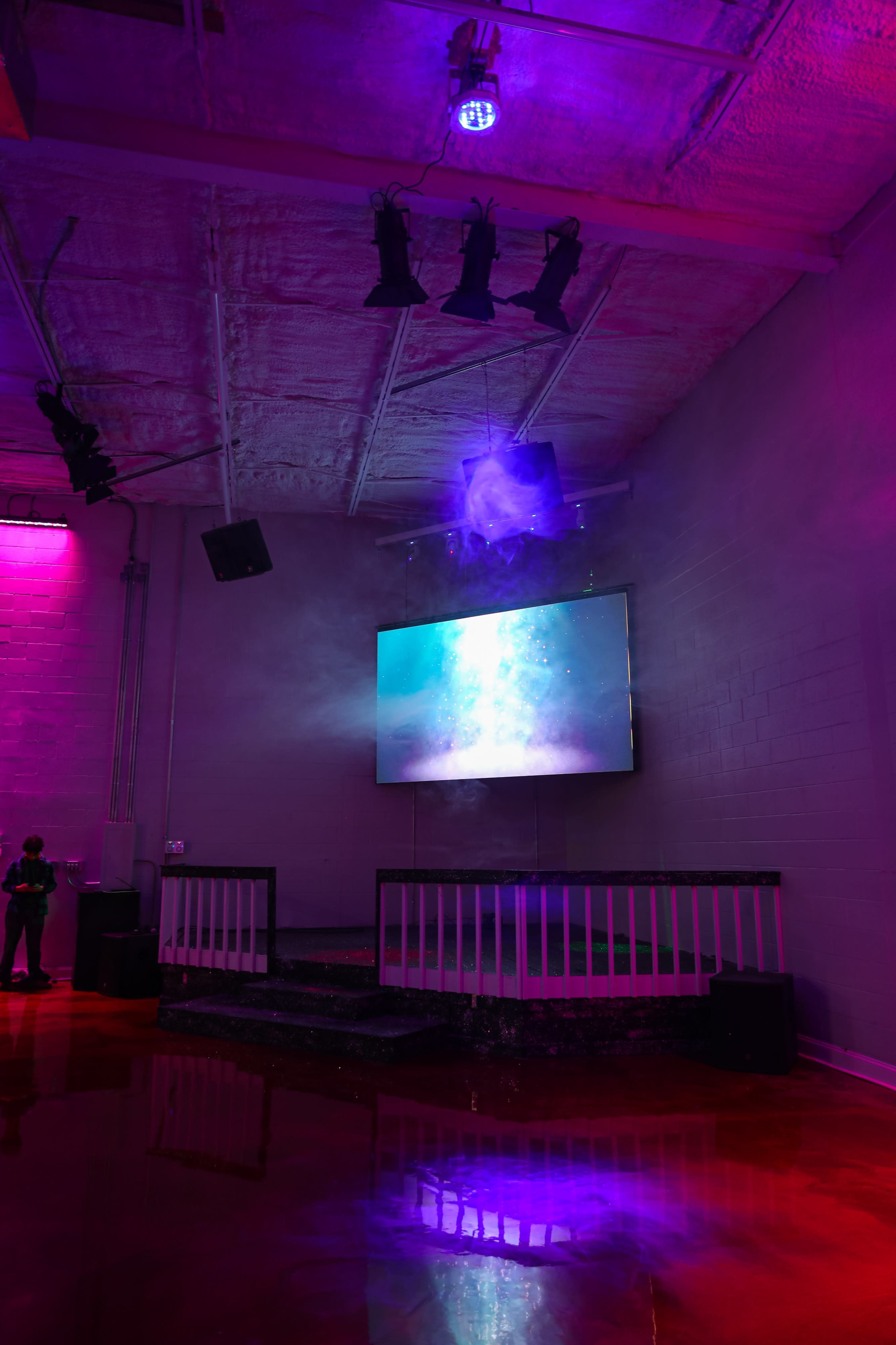 The image shows a dimly lit indoor venue with purple and blue lighting, featuring a large screen mounted on the wall and a mist effect around the stage area.