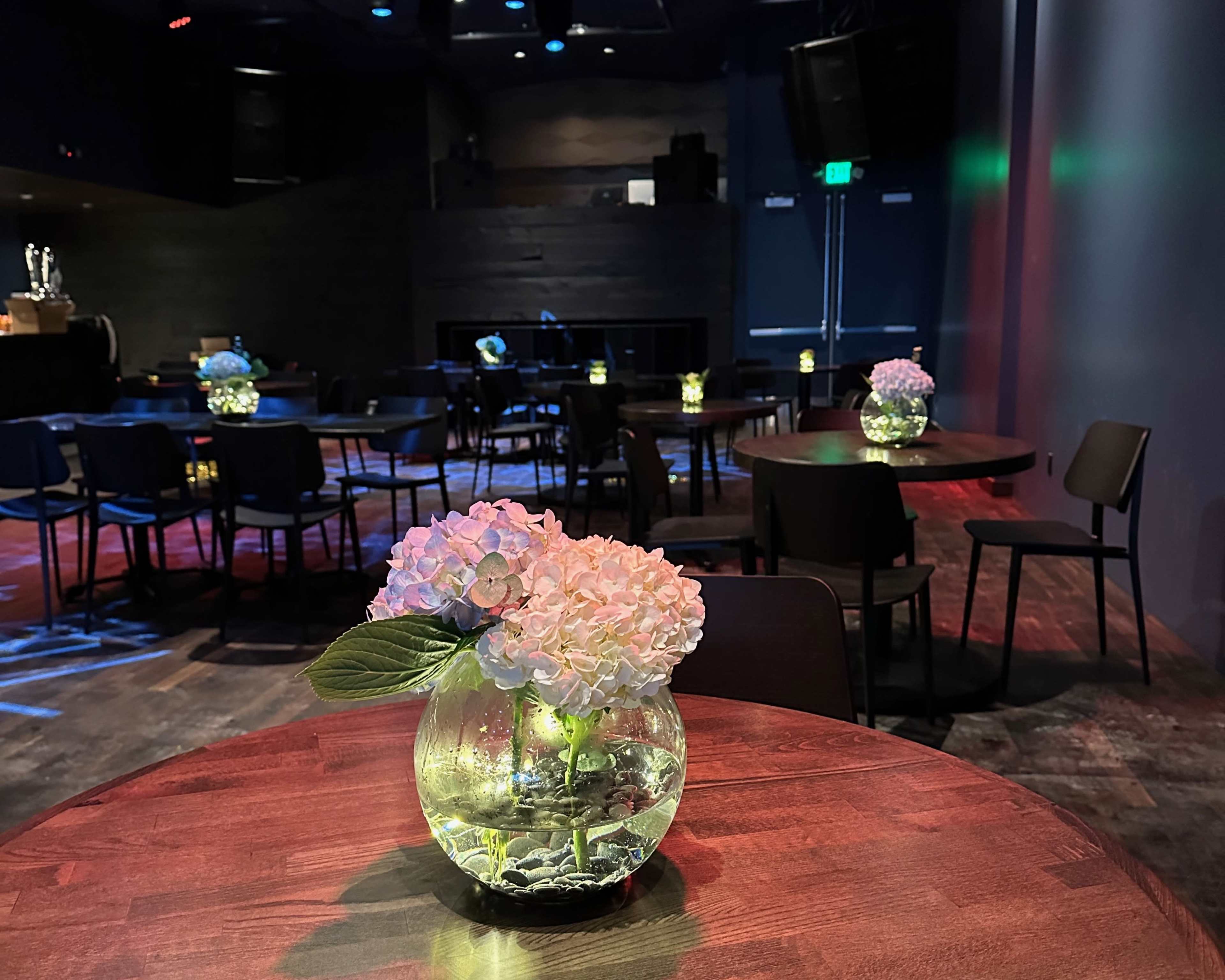 The image shows a dimly lit venue with several tables, each featuring a glass vase containing flowers, set against a backdrop of colorful lighting.