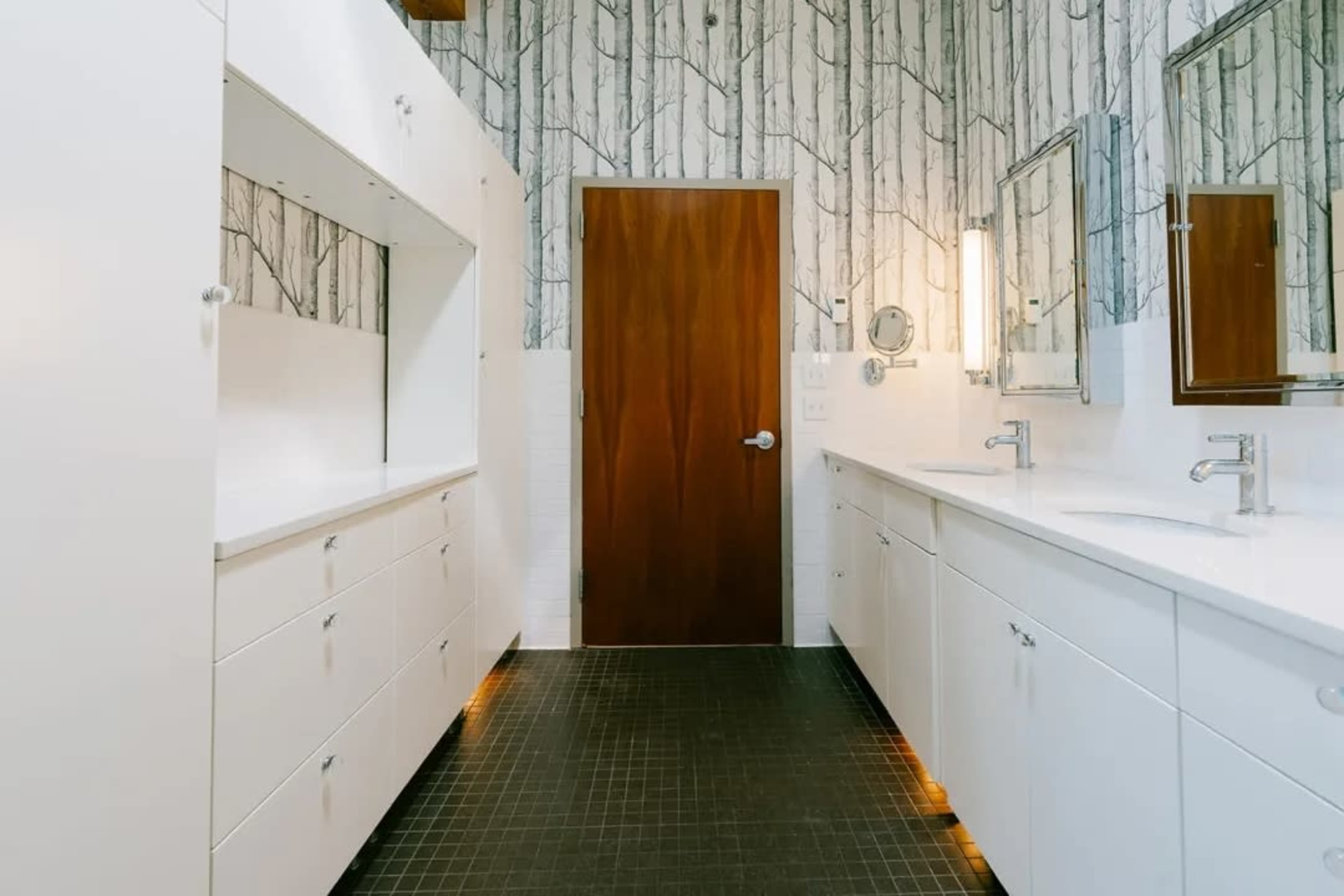 A modern bathroom features white cabinetry, dual sinks, and a door against a backdrop of tree-patterned wallpaper.