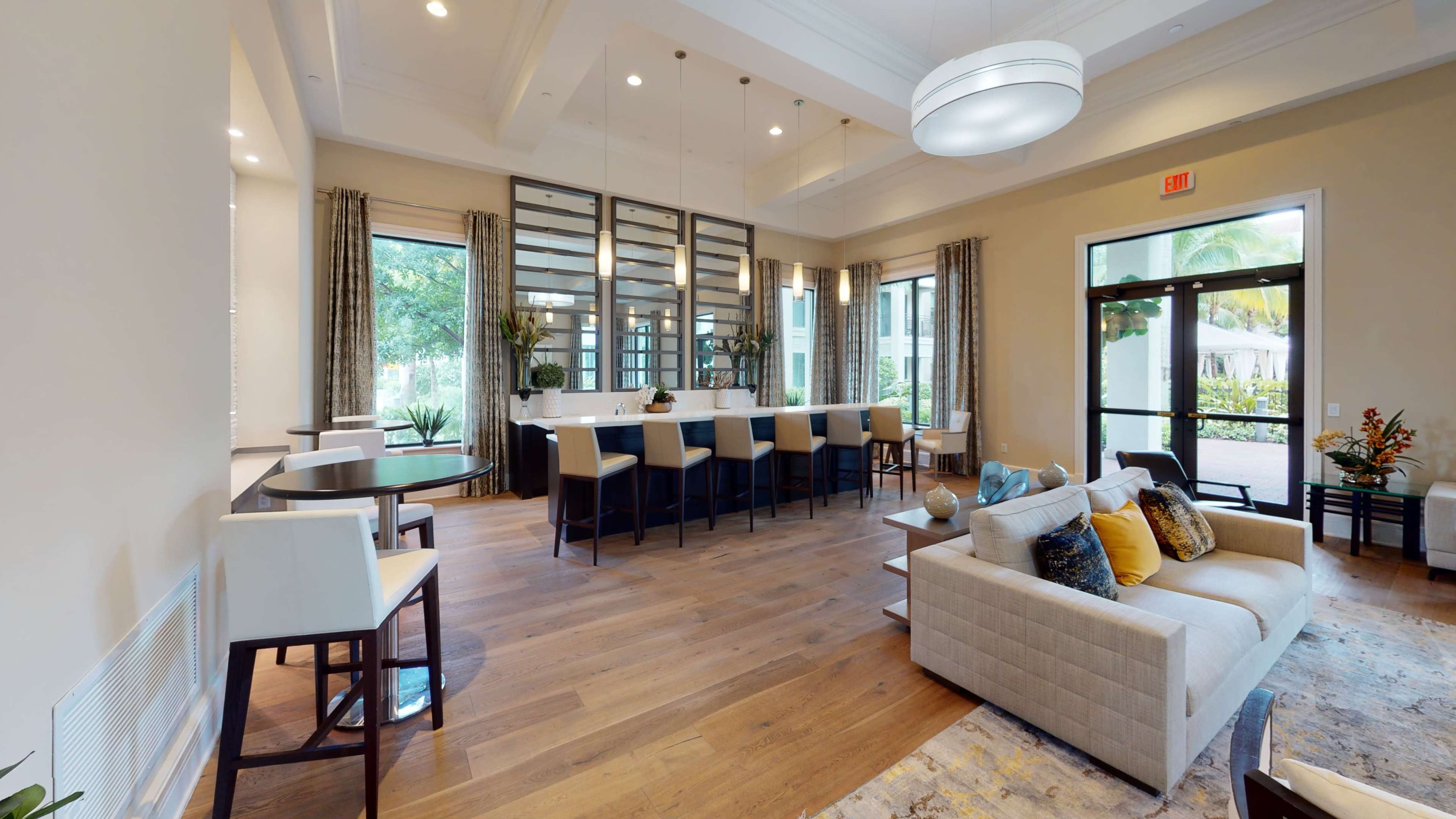 The image shows a modern lounge area with a long bar, seating at tables, and large windows letting in natural light.