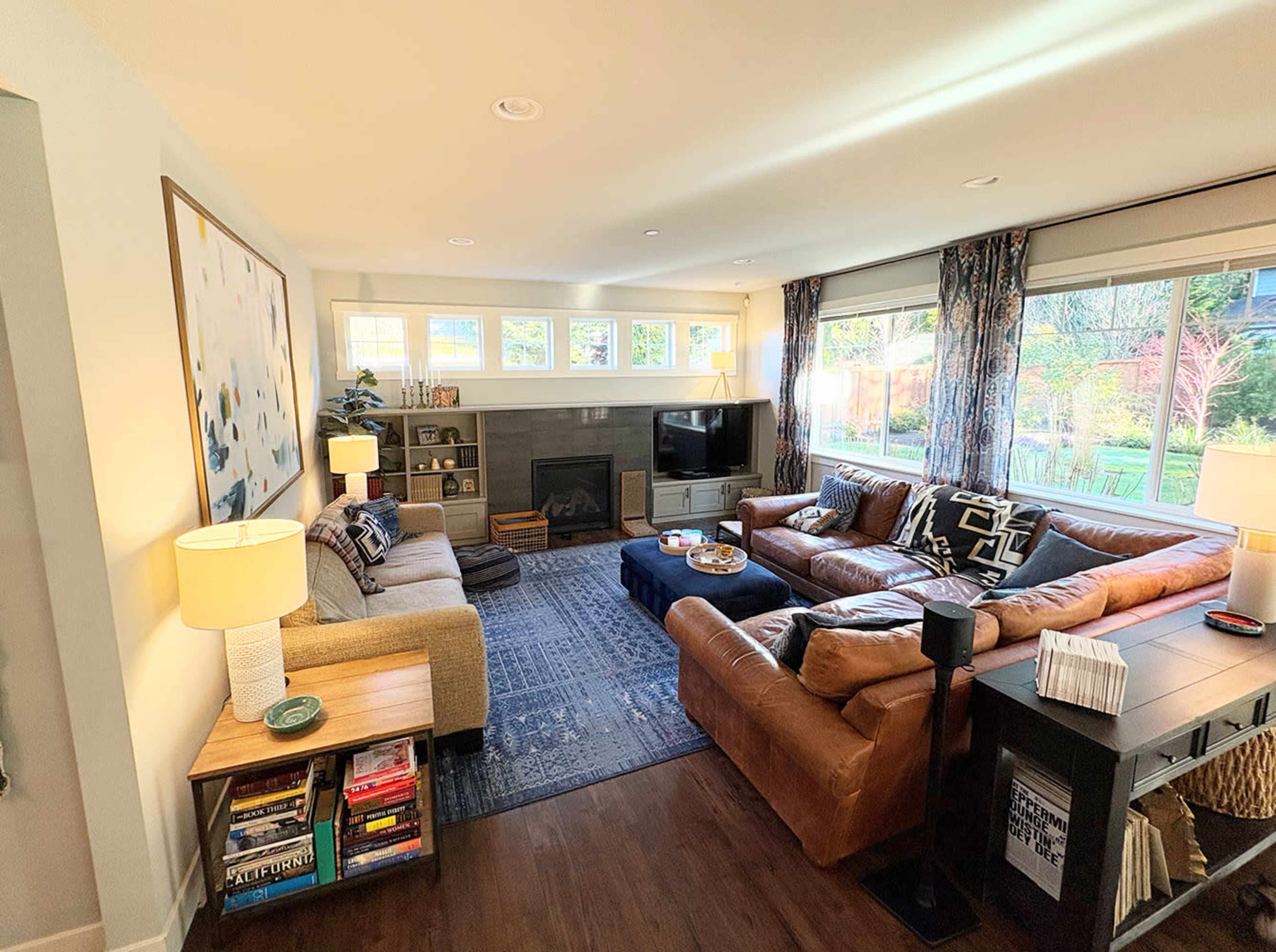 The image shows a spacious living room with two large sofas, a coffee table in the center, bookshelves on one side, and large windows letting in natural light.
