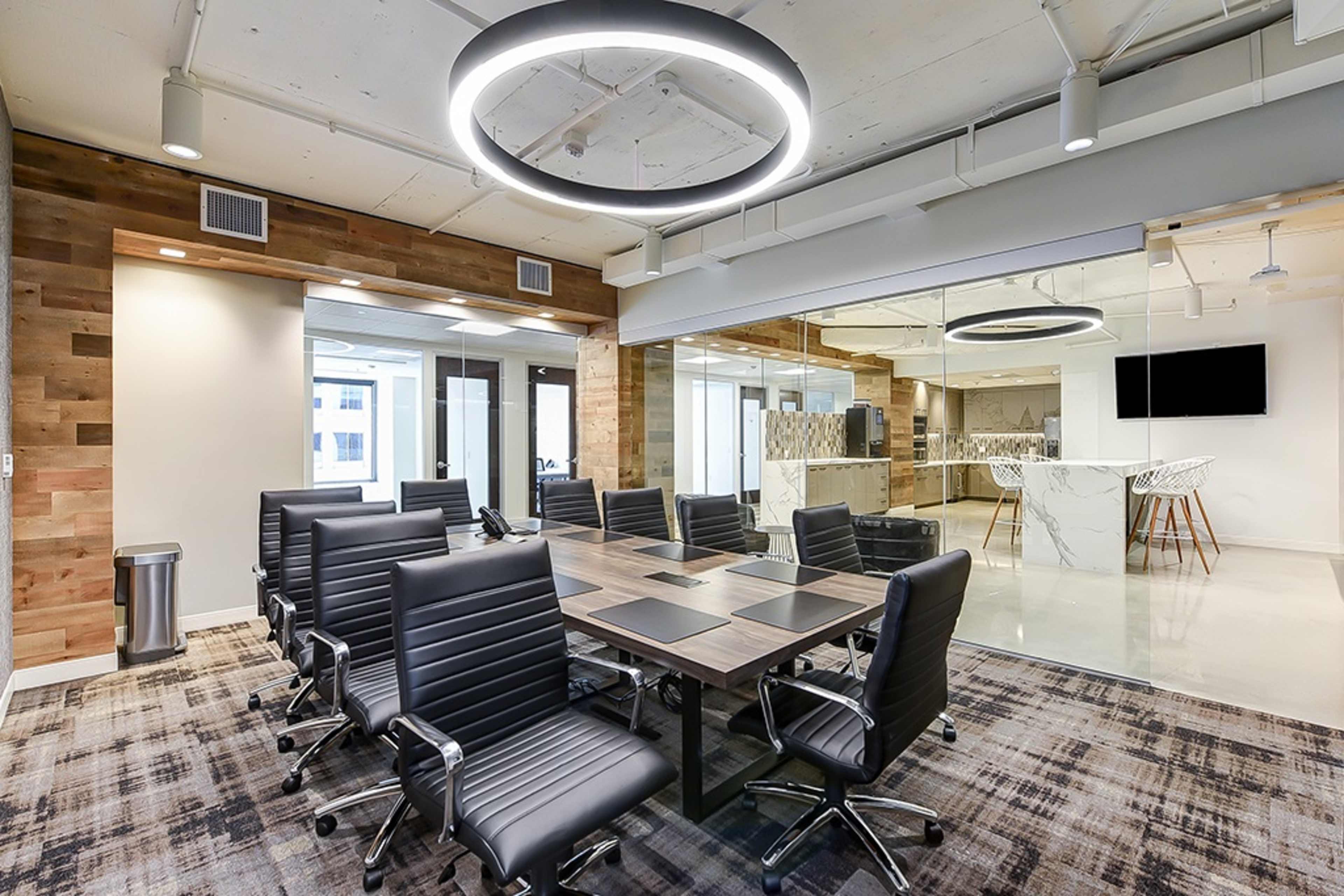 A modern conference room features a large table surrounded by black leather chairs, with a circular overhead light and a glass wall leading to a kitchen area.