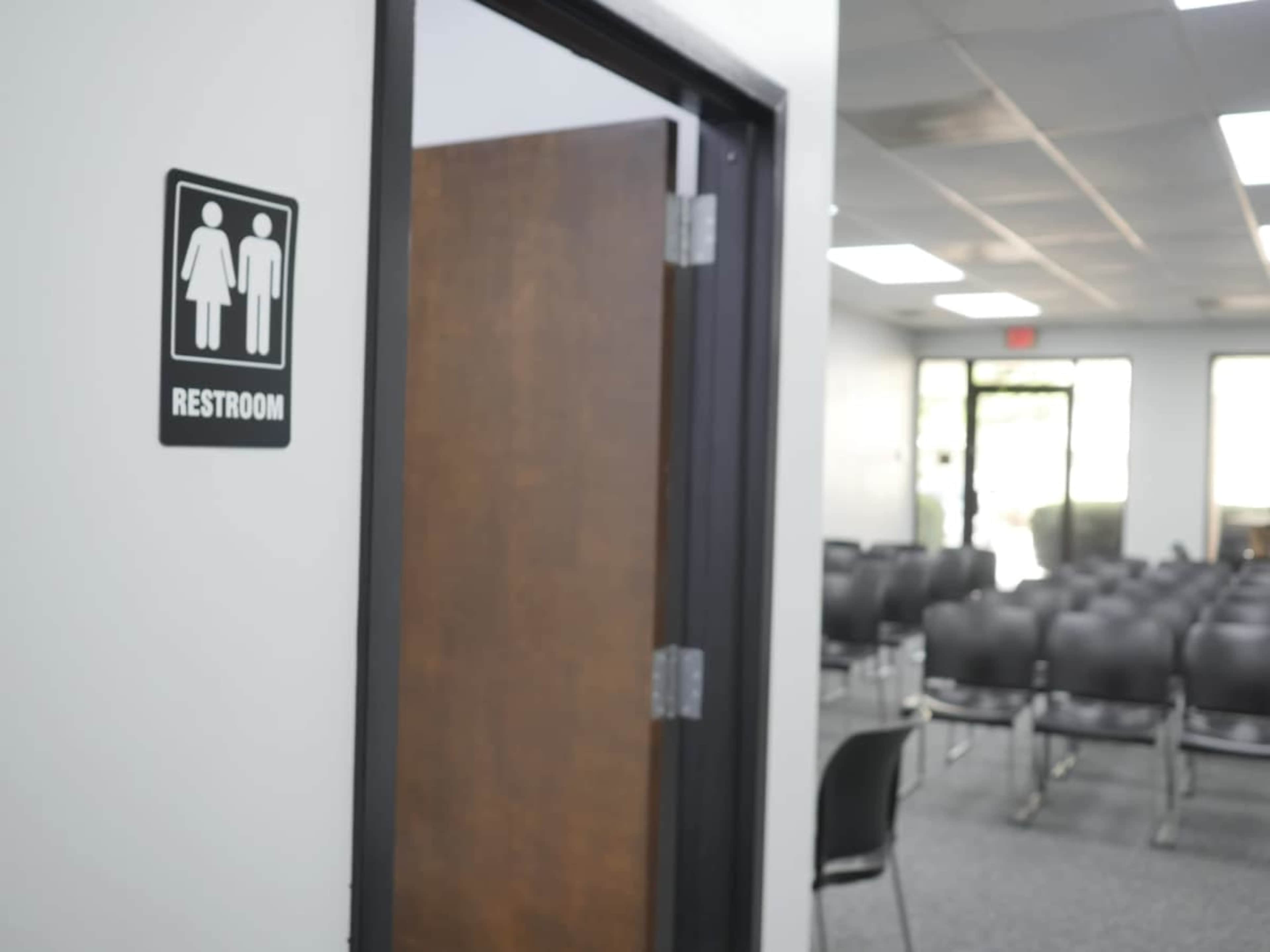 The image shows a restroom sign on a wall next to an open door leading to a waiting area with rows of chairs.