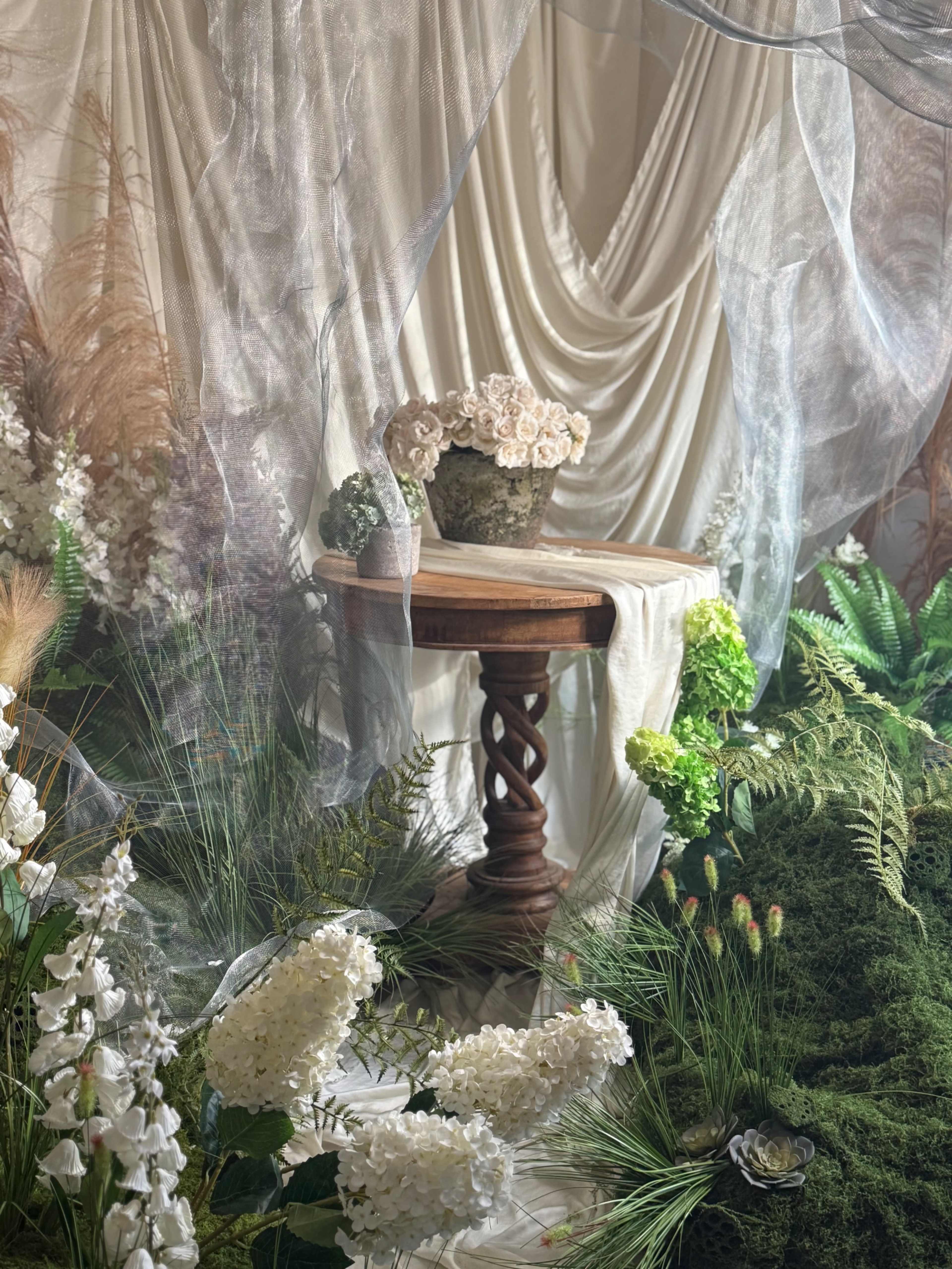 A wooden table adorned with a floral arrangement is set within a backdrop of draped fabric and greenery.