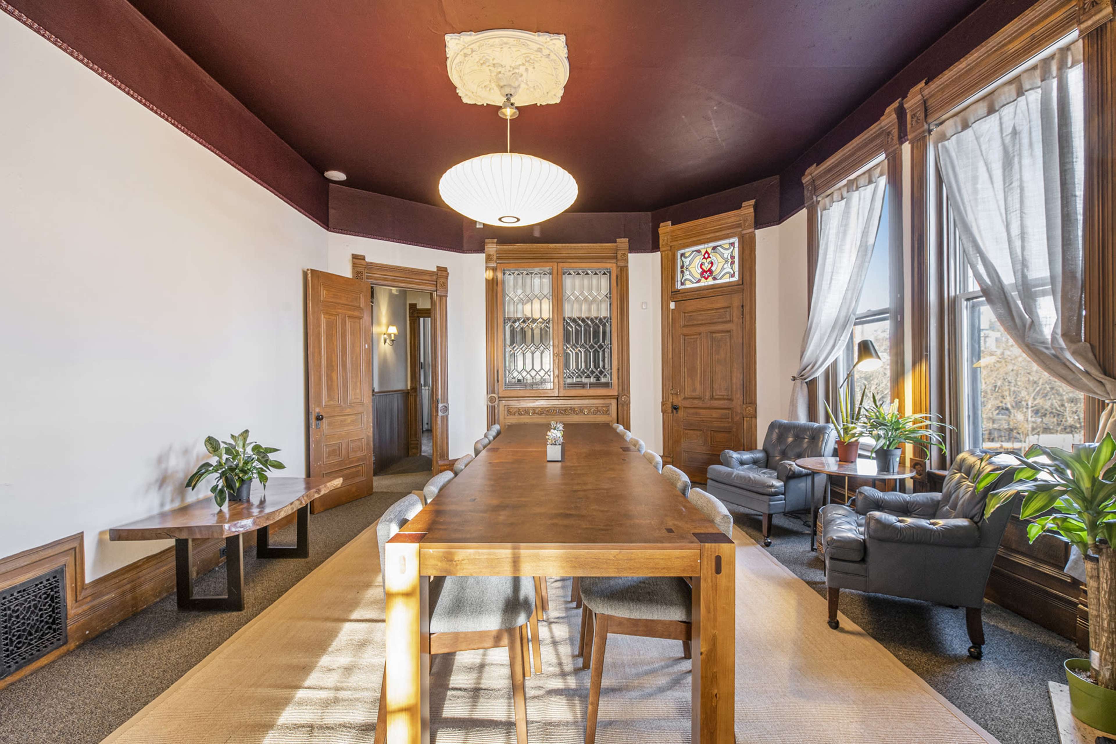 The image shows a spacious room with a wooden dining table, surrounded by chairs, and large windows that let in natural light, along with potted plants and decorative elements.