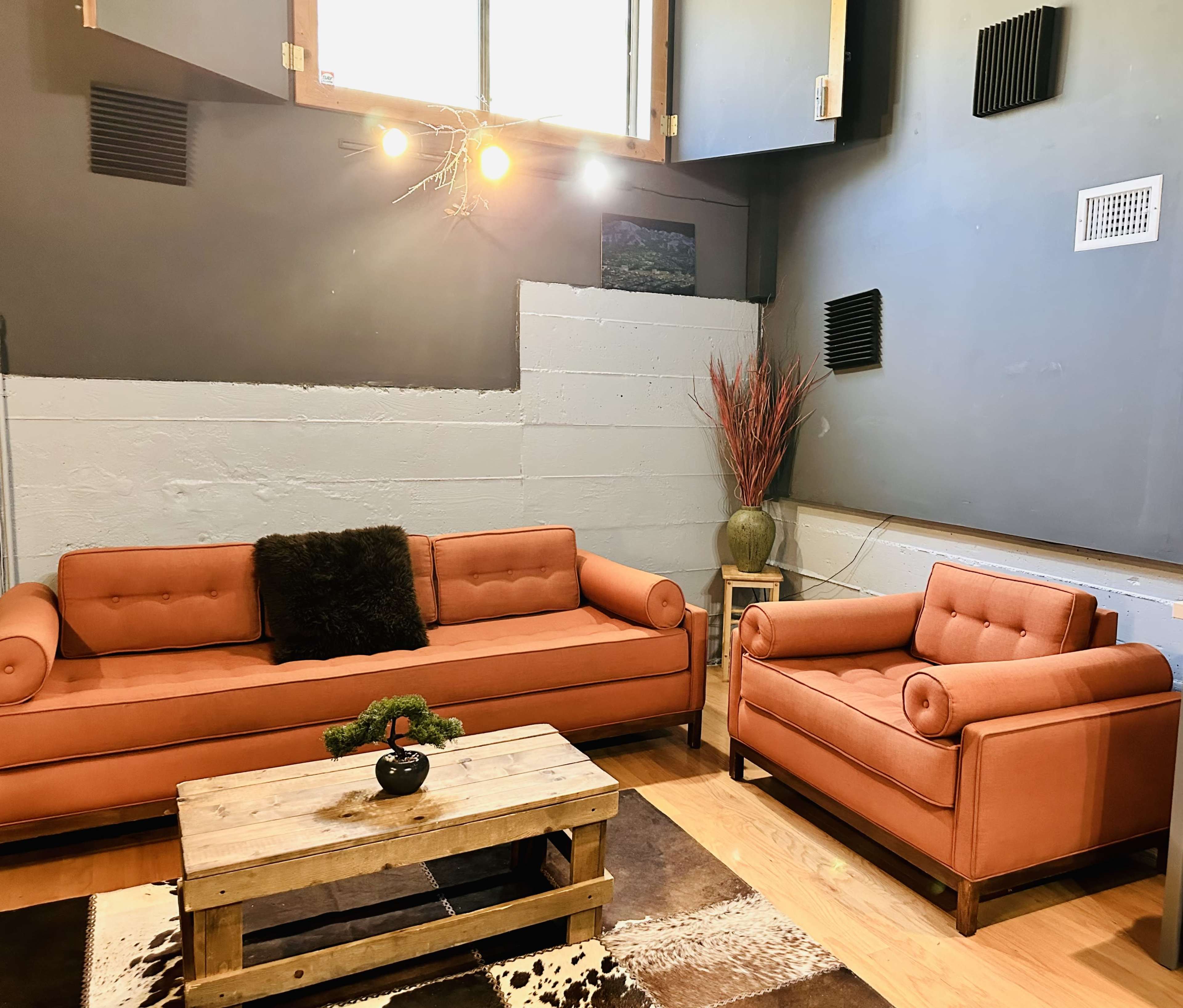 The image shows a modern living room featuring an orange sofa and armchair, a wooden coffee table, and decorative plants against a gray wall.