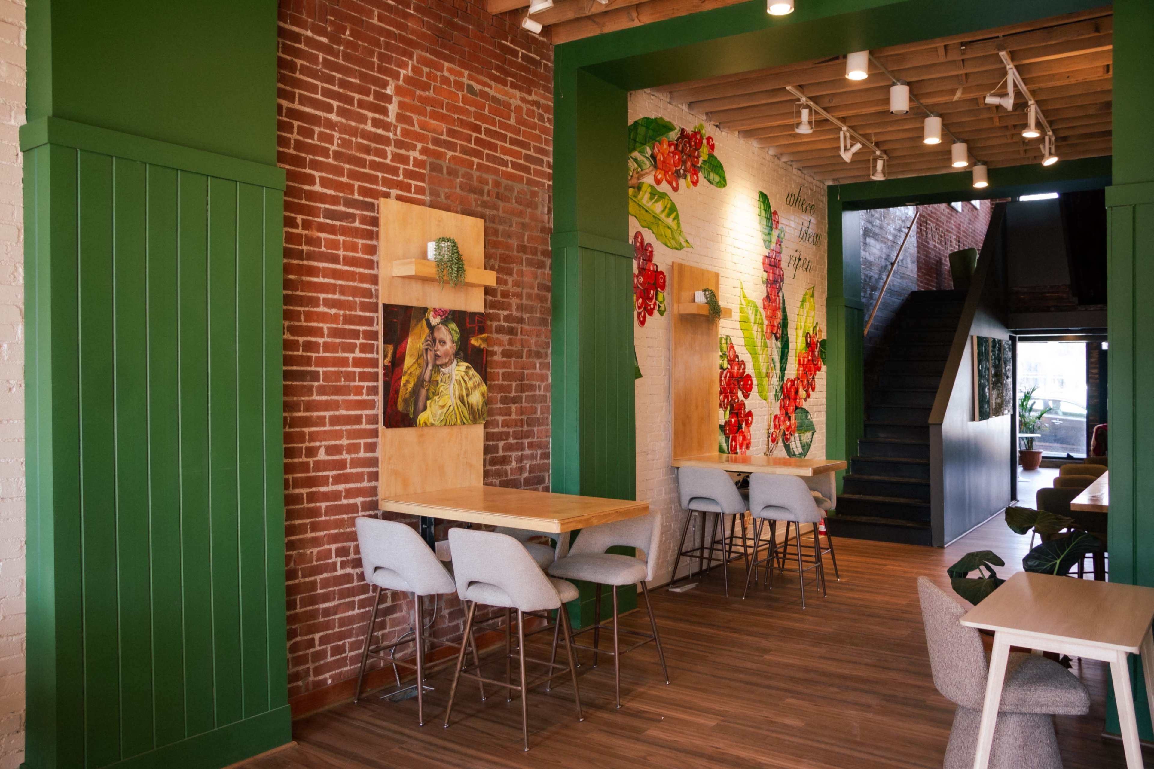 The image shows a modern cafe interior featuring exposed brick walls, green accents, and artwork of plants, with seating arrangements that include light-colored chairs and wooden tables.