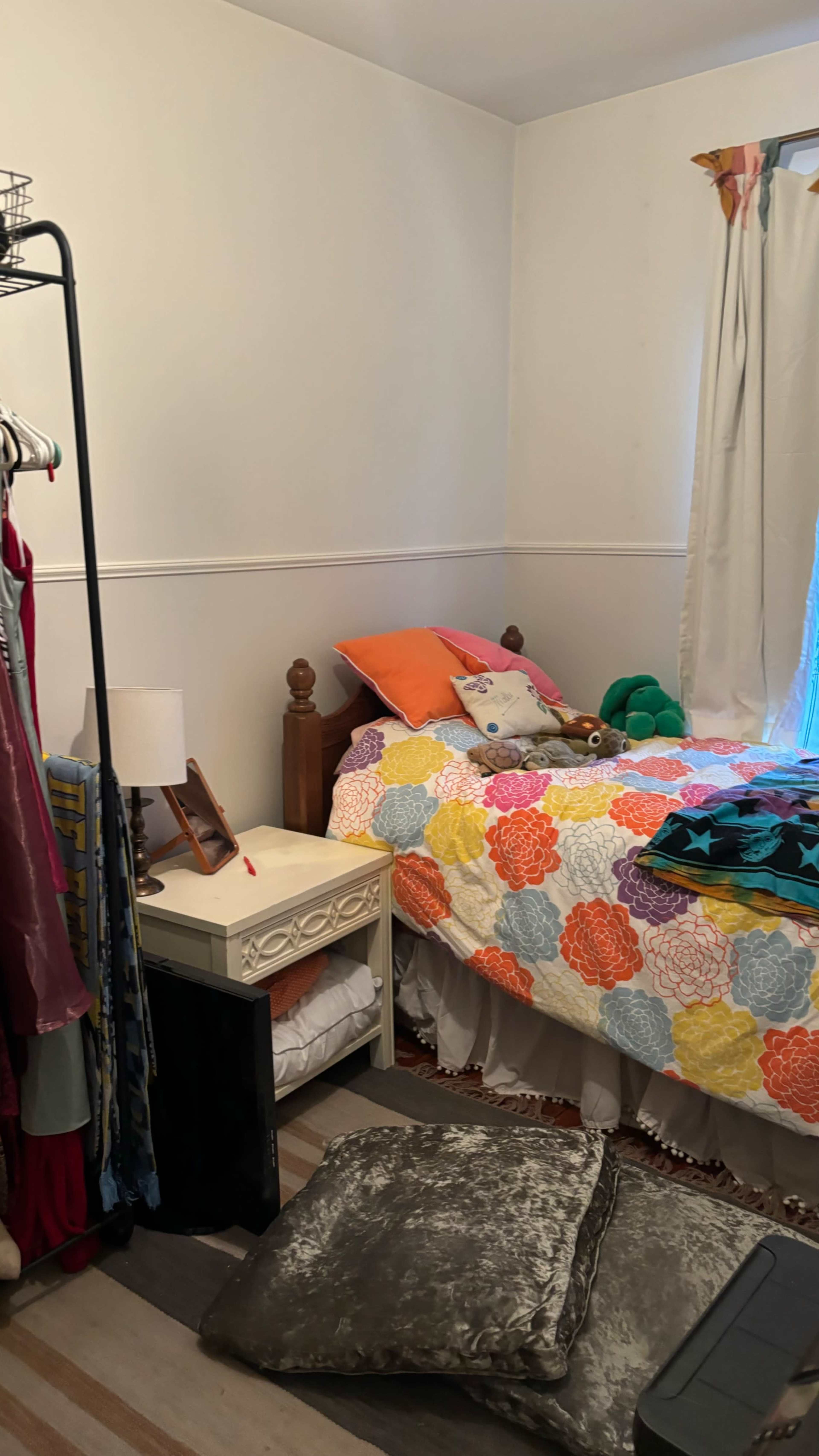 A brightly colored bedroom features a neatly made bed with a floral quilt, surrounded by a clothing rack and decorated with plush toys.