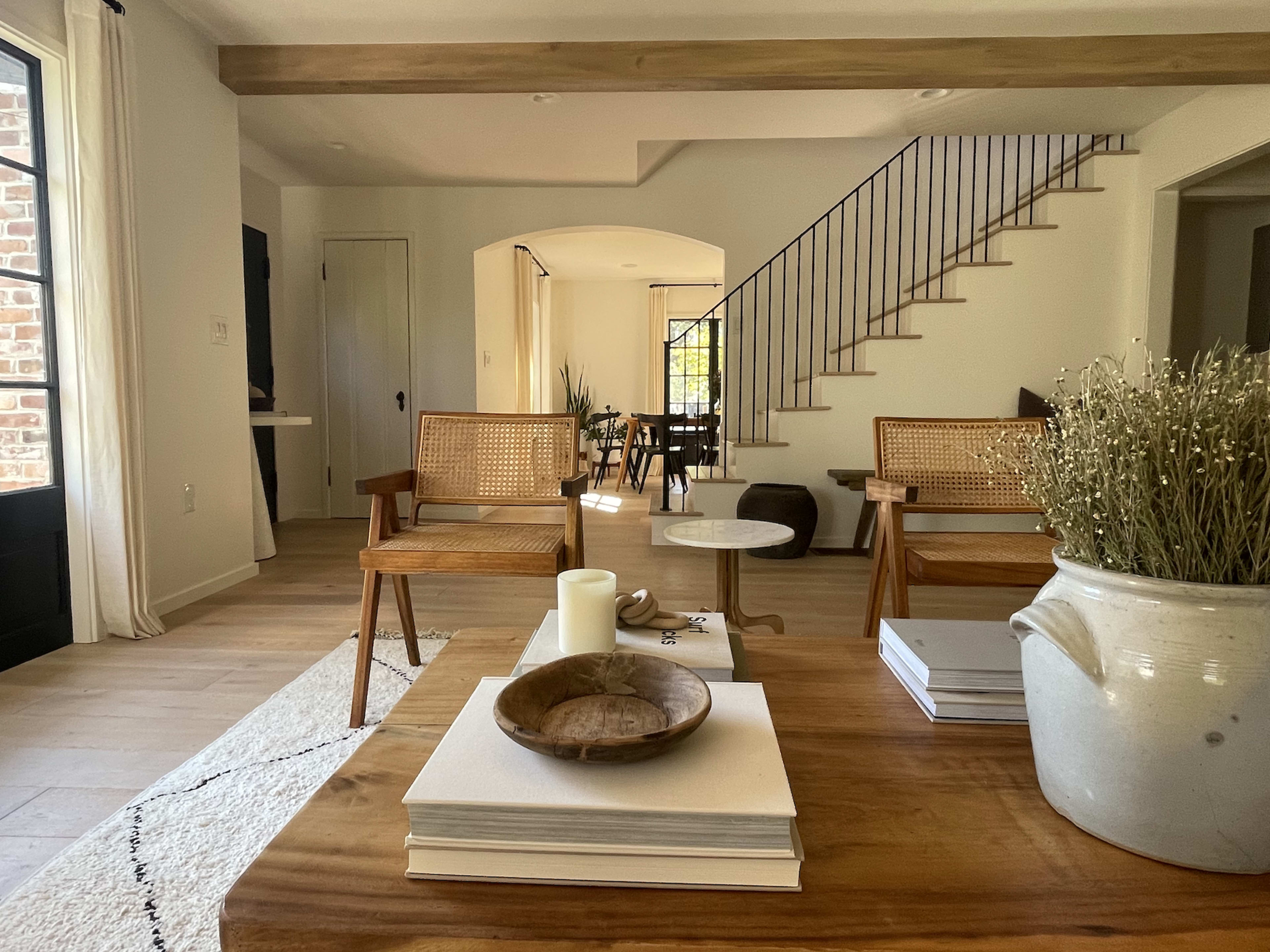 The image depicts a light-filled living area featuring wooden furniture, a round coffee table, and a staircase in the background.