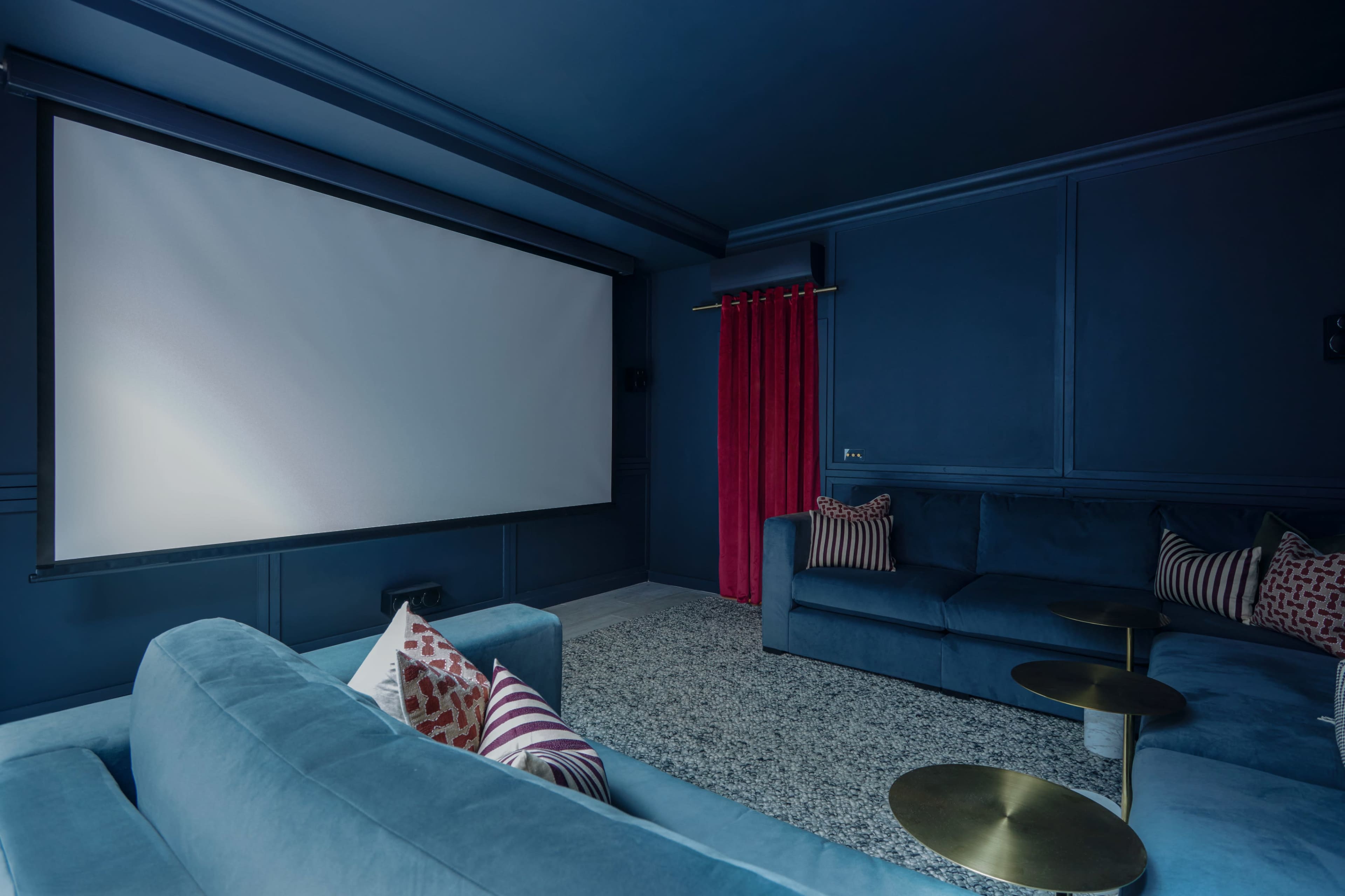 A home theater room features a large white projection screen, deep blue walls, and plush seating with decorative cushions.