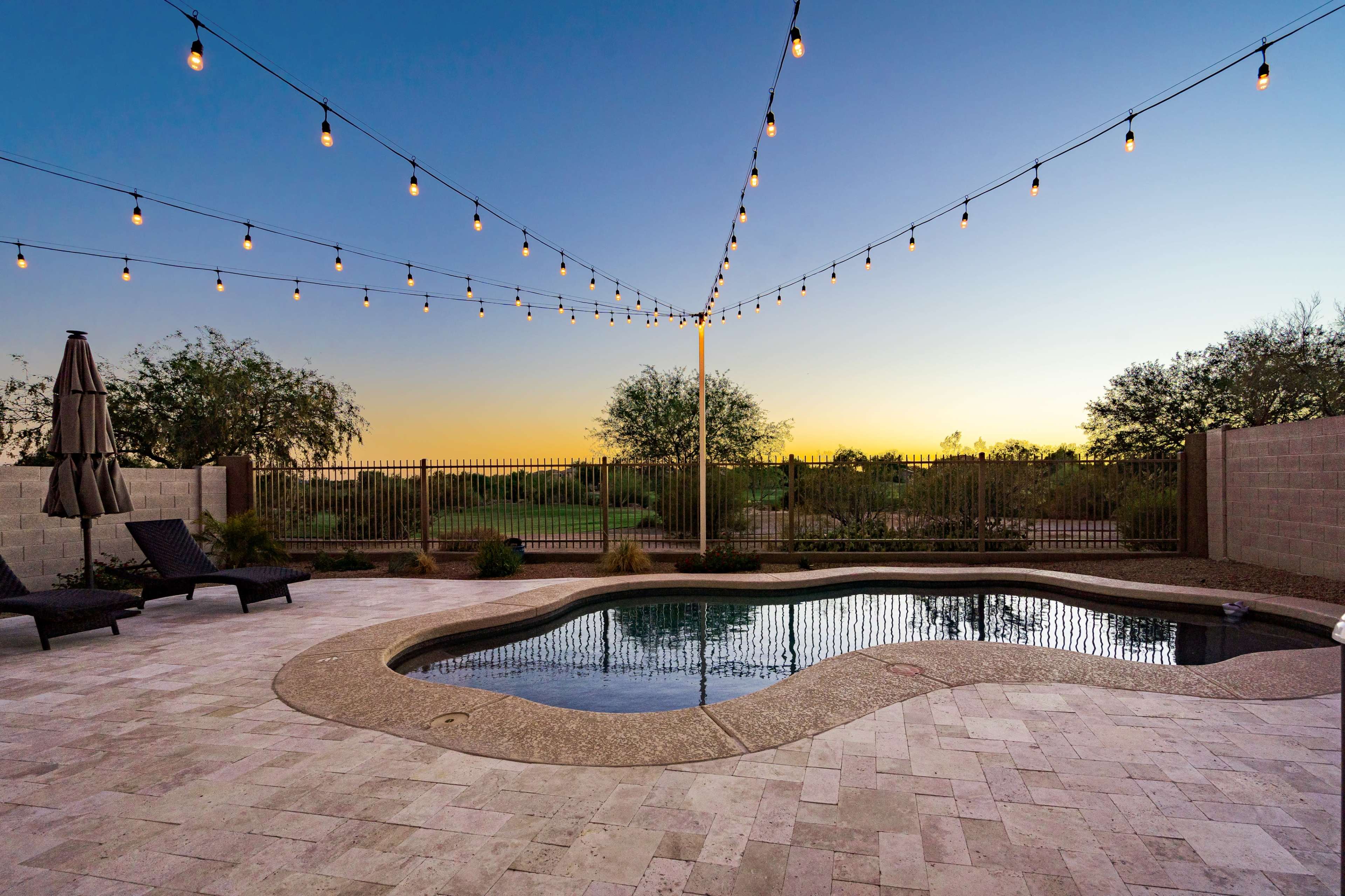 A swimming pool with a curved edge is surrounded by patio furniture and illuminated by string lights at dusk.