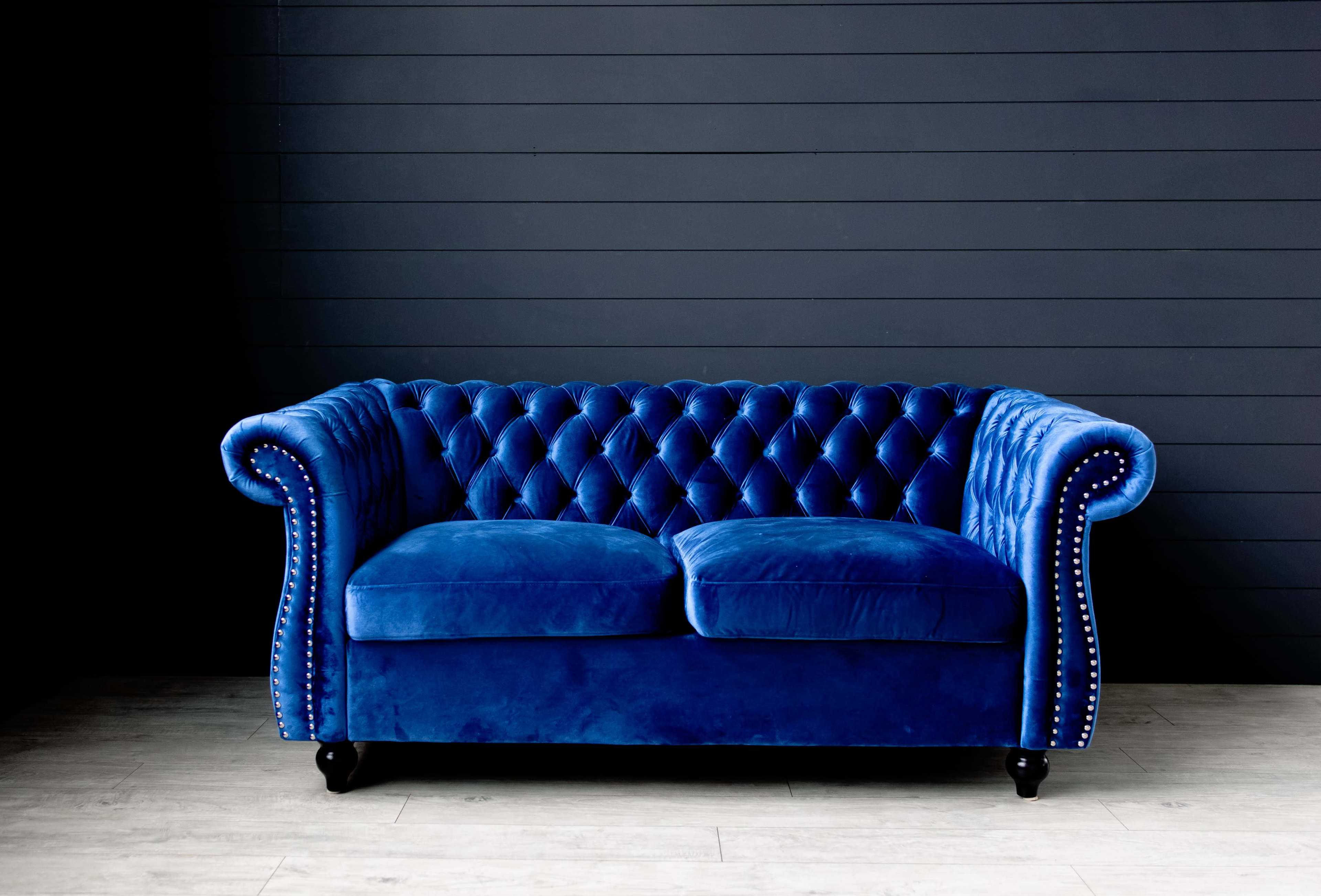 A blue velvet chesterfield sofa with tufted upholstery and wooden legs is positioned against a dark wall.