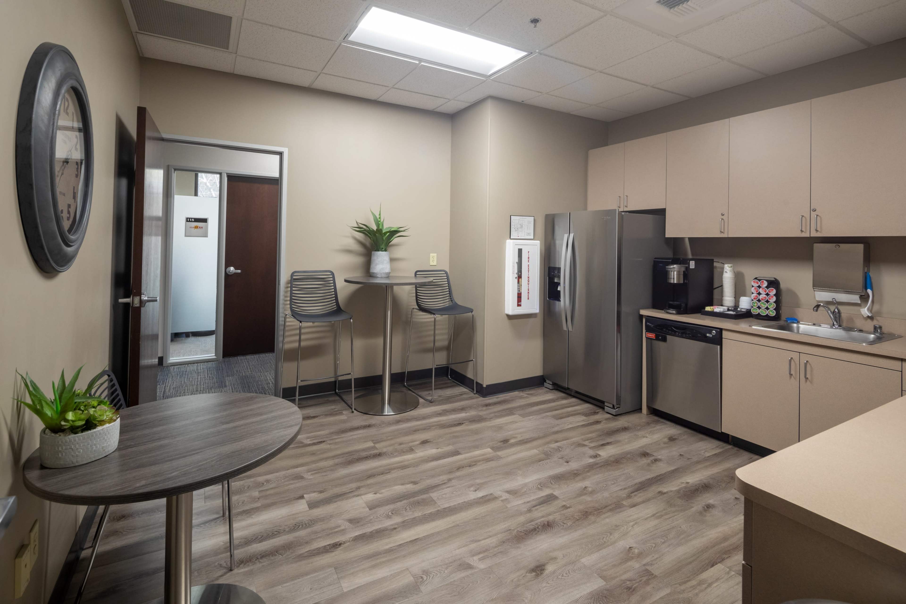 The image shows a modern break room with a kitchenette, including a refrigerator, coffee maker, and two high tables.